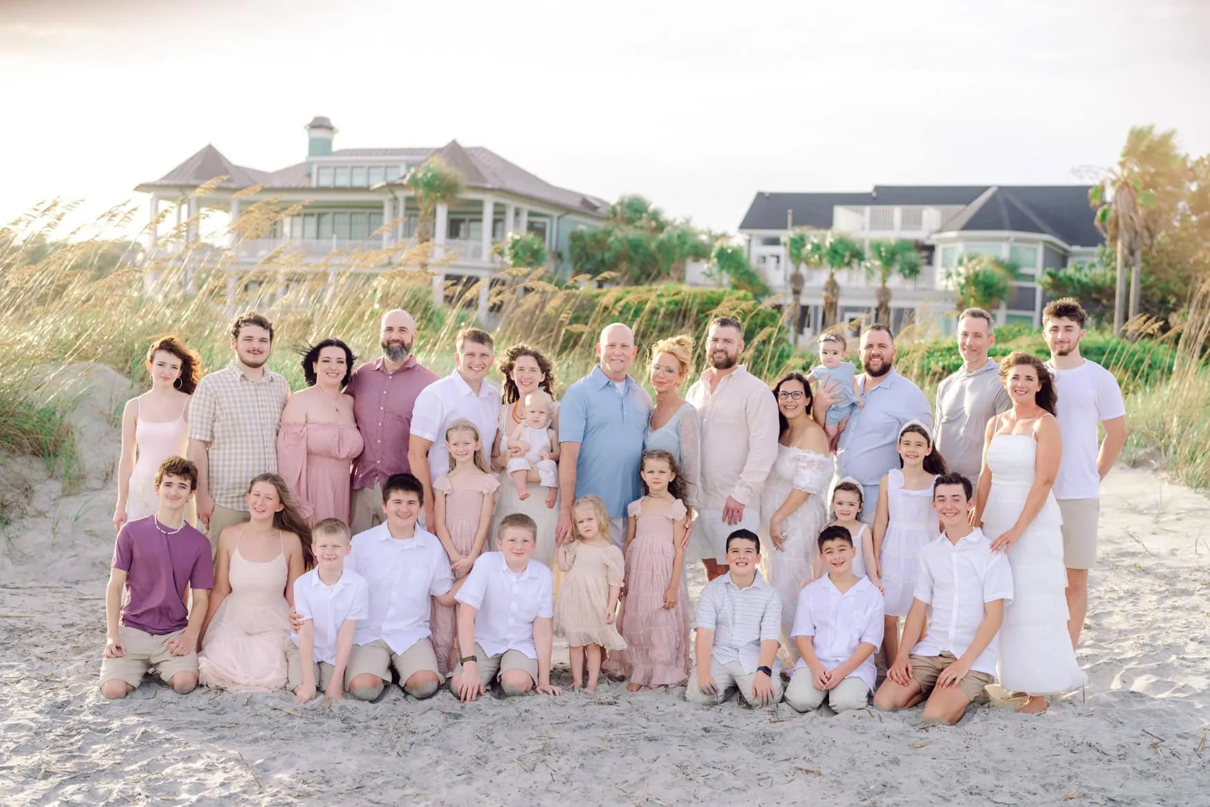 29 person extended family portrait on the beach at Wild Dunes Resort in Isle of Palms near Charleston, South Carolina. Multi generational family posing together at sunset with sea oats, beach homes, and ocean dunes in the background during a professional family photography session.