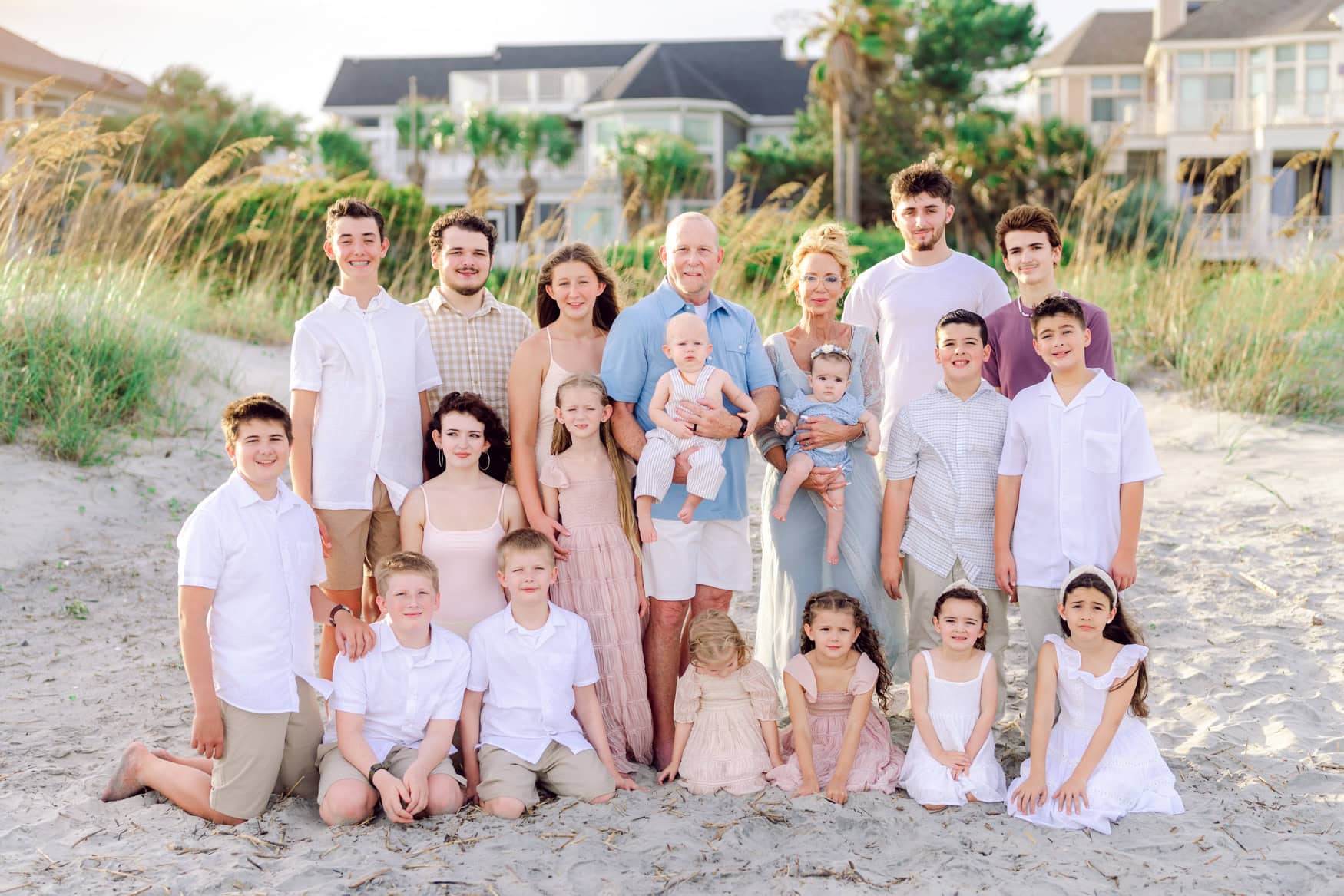 Grandparents surrounded by their grandchildren during a family beach portrait at Wild Dunes Resort in Isle of Palms near Charleston, South Carolina. Multi generational family photography session on the beach with sea oats, sand dunes, and coastal homes in the background at sunset.