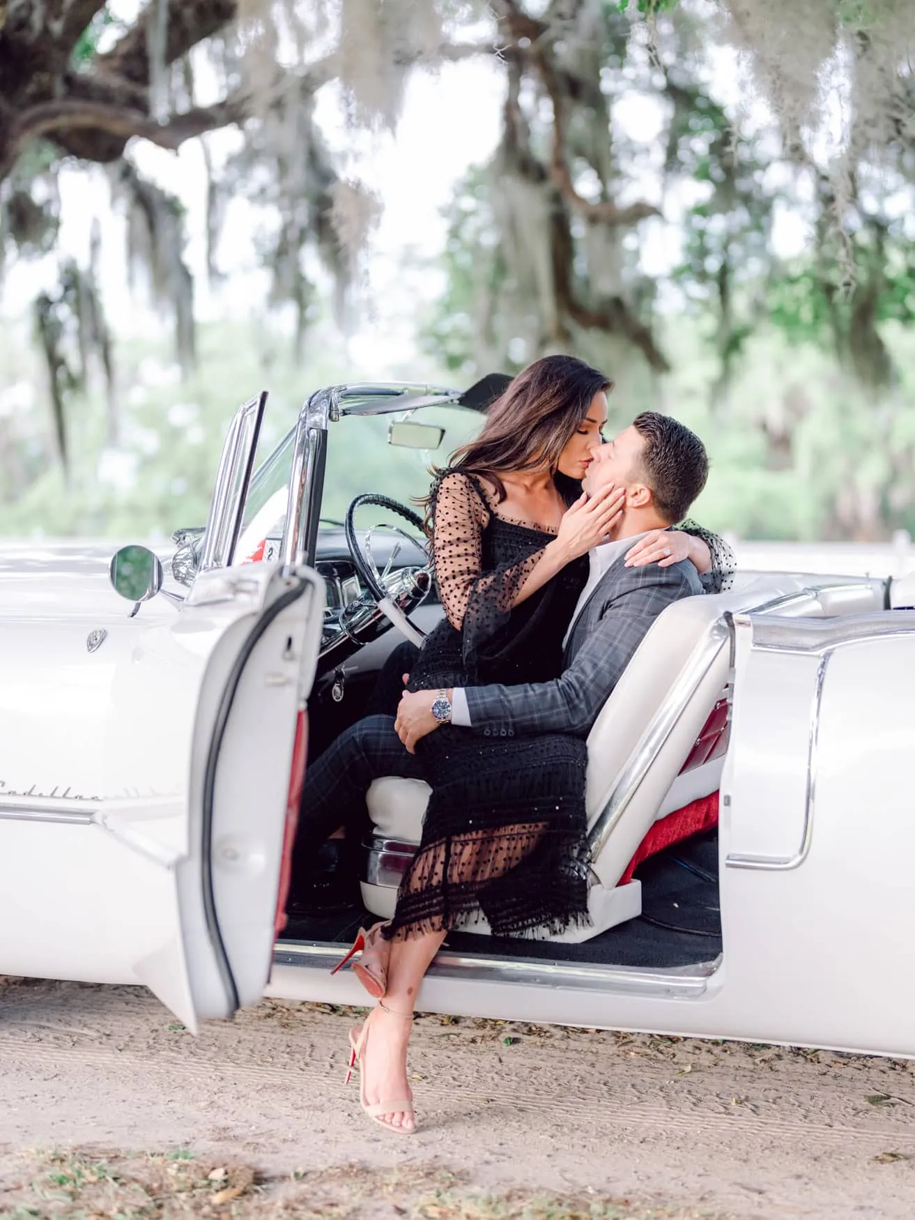 Couple in a black dress and gray suit seated in a white vintage convertible under Spanish moss