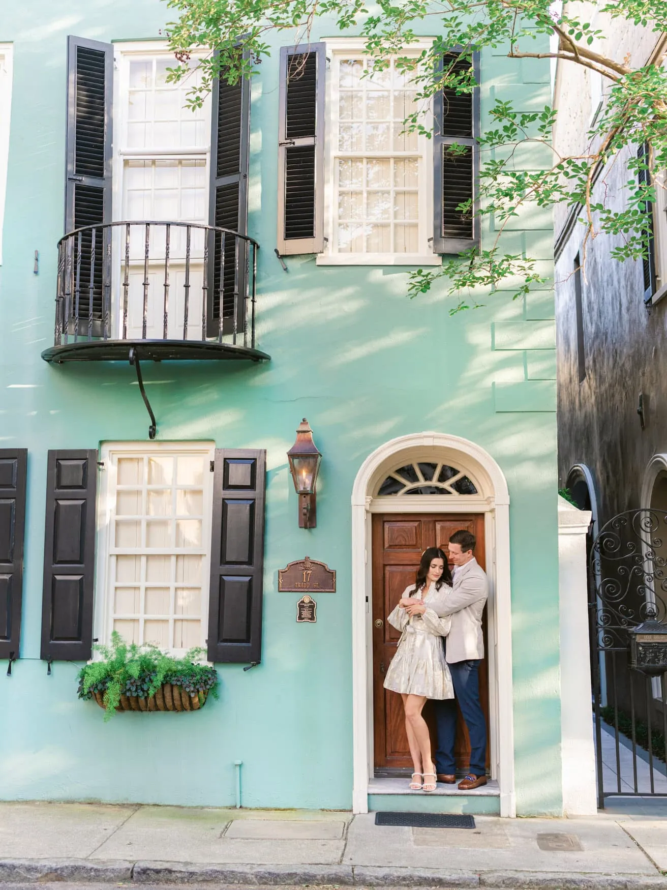 Couple in a metallic short dress and light blazer beside a turquoise historic home in Charleston SC