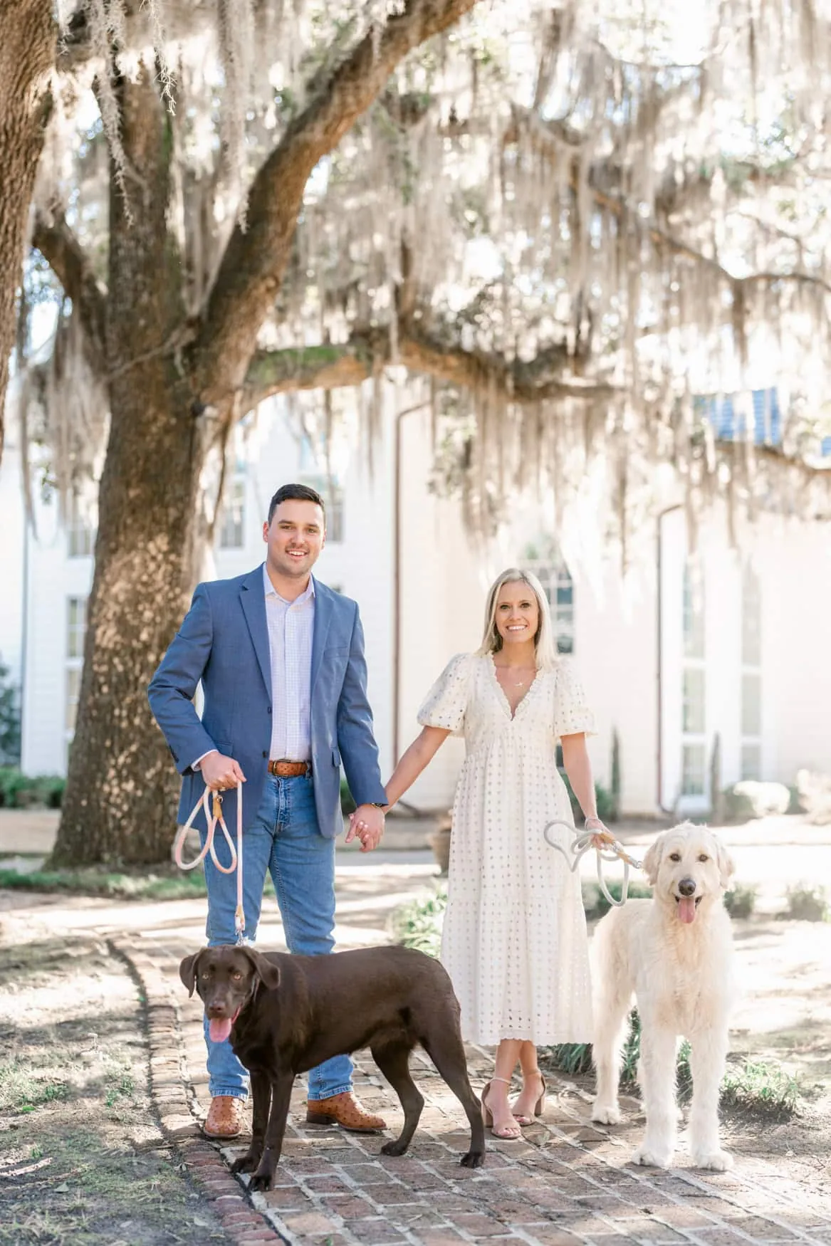 Couple in a white midi dress and blue blazer walking with their dogs under Spanish moss in Charleston