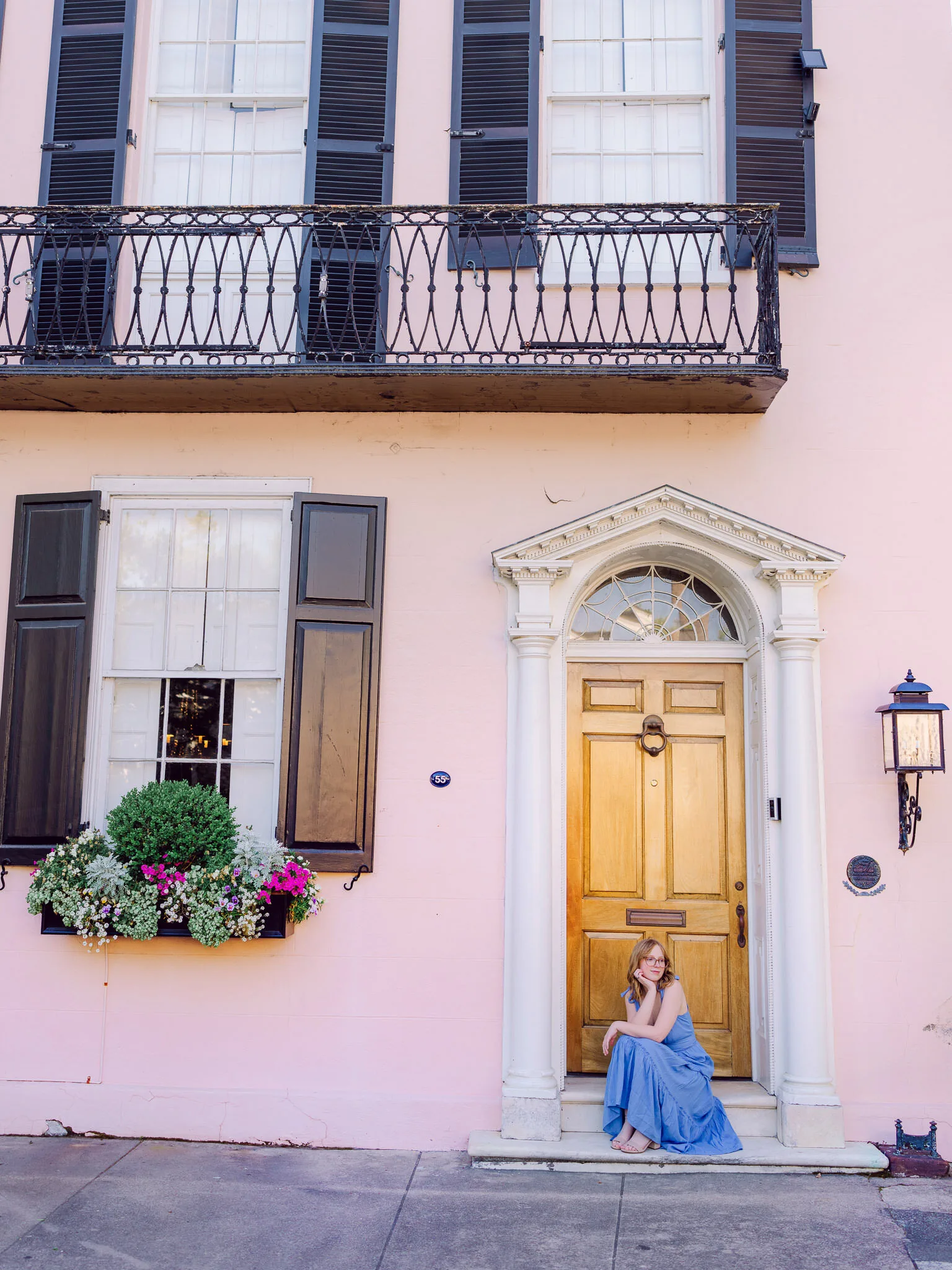 Senior girl in a blue dress sitting at the yellow door of a pink Charleston house with a flower box balcony
