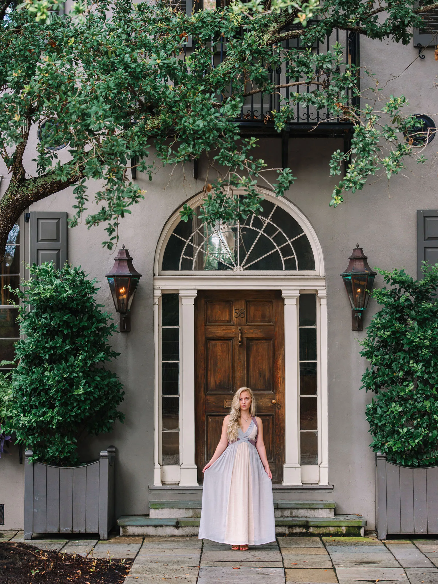 Senior girl in a pale lavender dress standing at an arched wooden doorway of a Charleston townhouse with lanterns