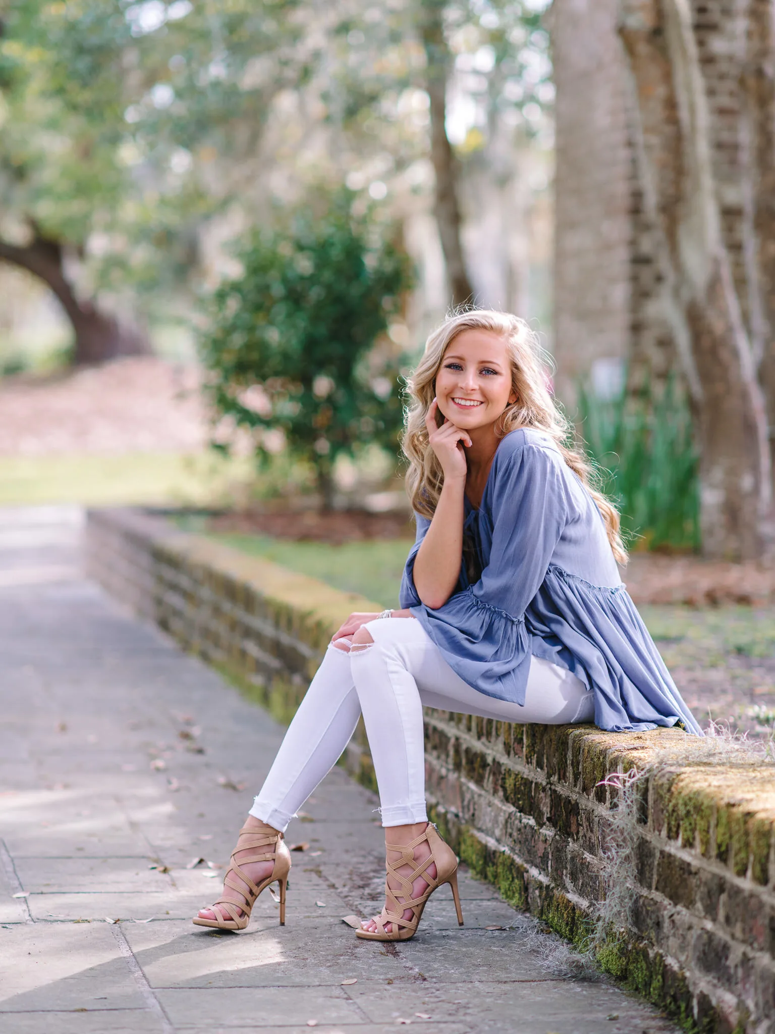Blonde senior girl in a periwinkle top and white jeans sitting on a mossy brick garden wall under live oaks in Charleston