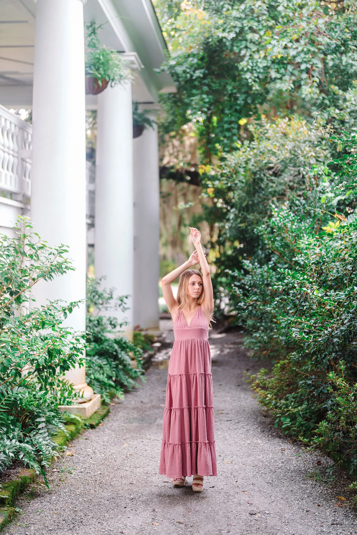 Senior girl in a rose maxi dress with arms raised on a garden path beside white columns in downtown Charleston