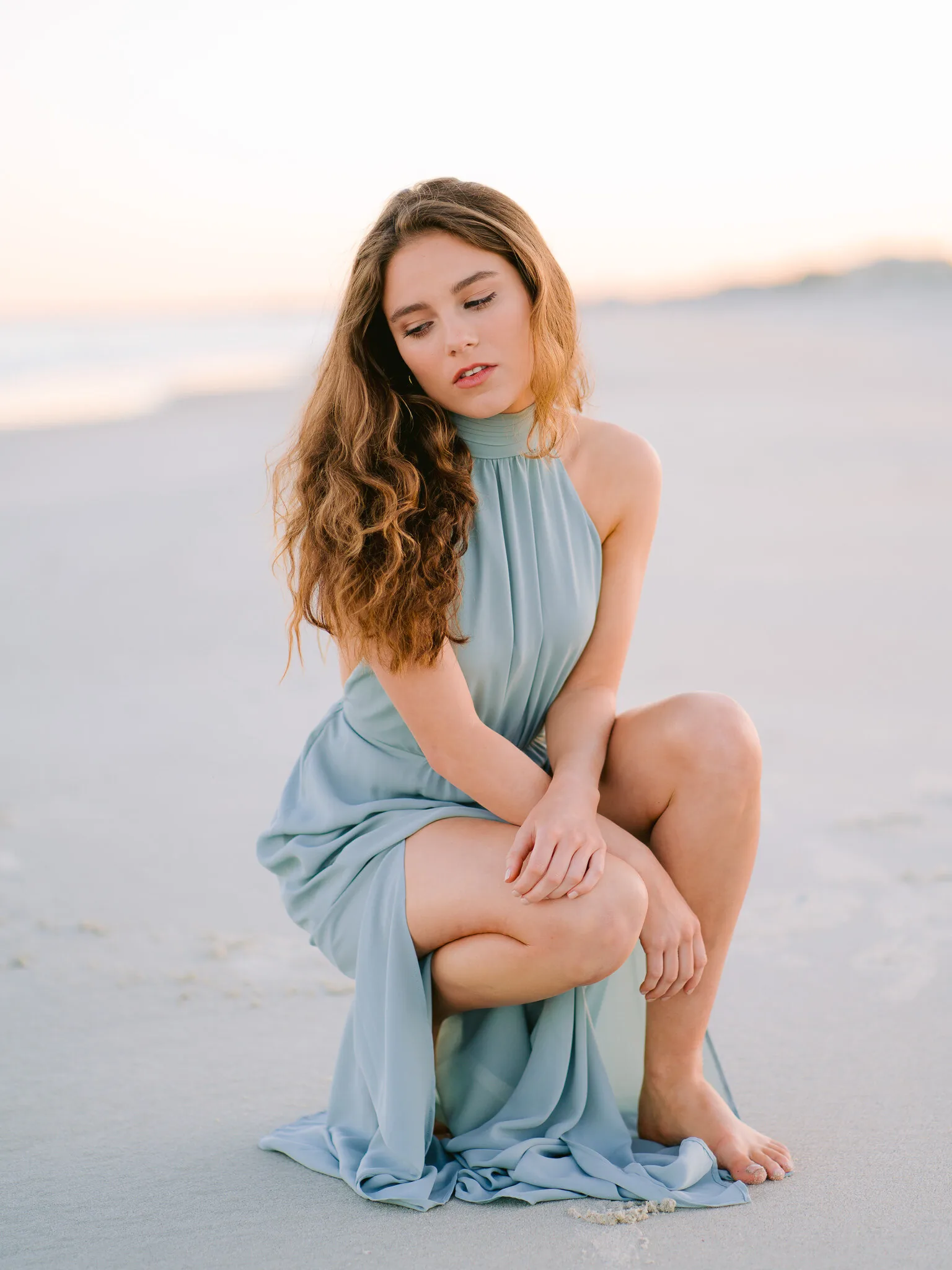 Senior girl in a sage green halter dress crouching on the sand at a soft pastel beach