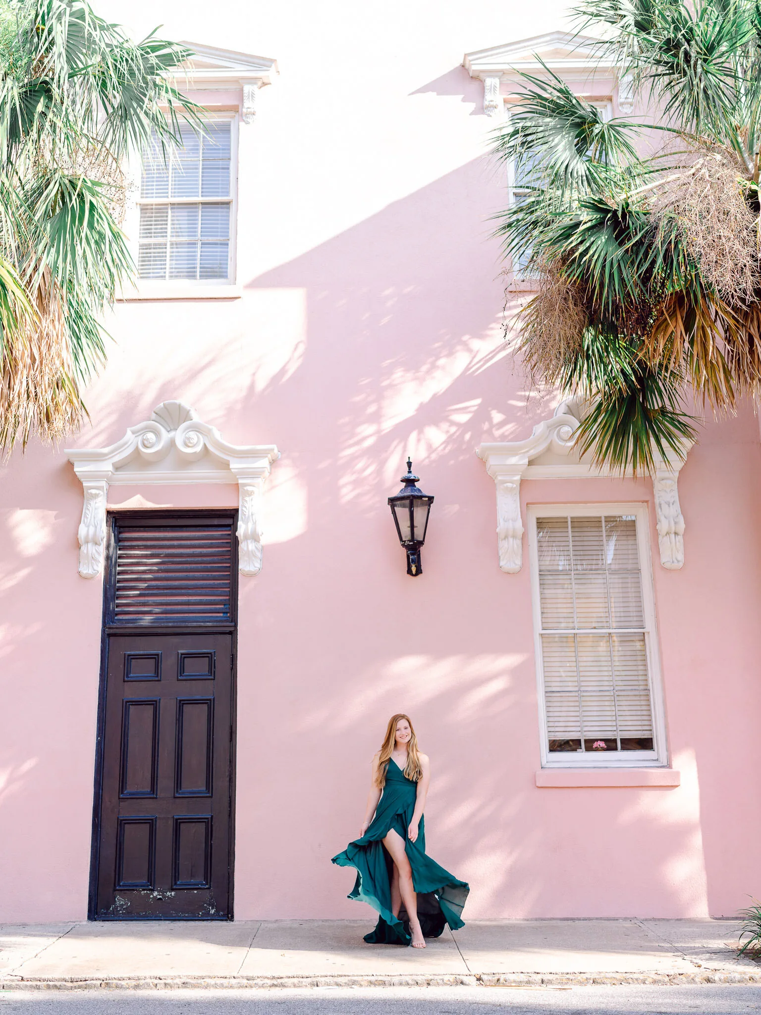 Wide view of a senior girl in an emerald dress standing beside a dark door on a pink Charleston house with palms