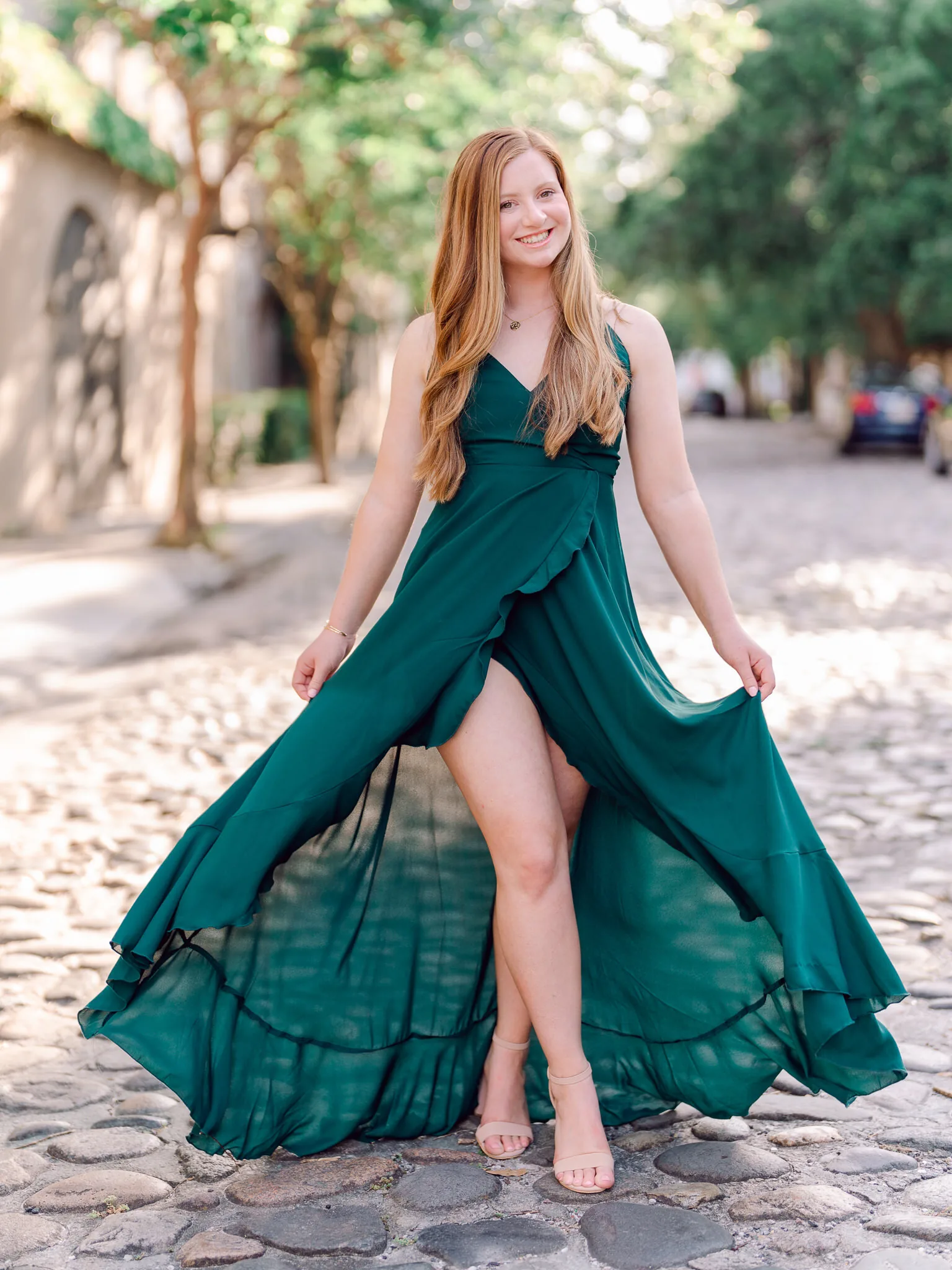 Senior girl in an emerald green wrap dress holding her skirt on a Charleston cobblestone street lined with trees