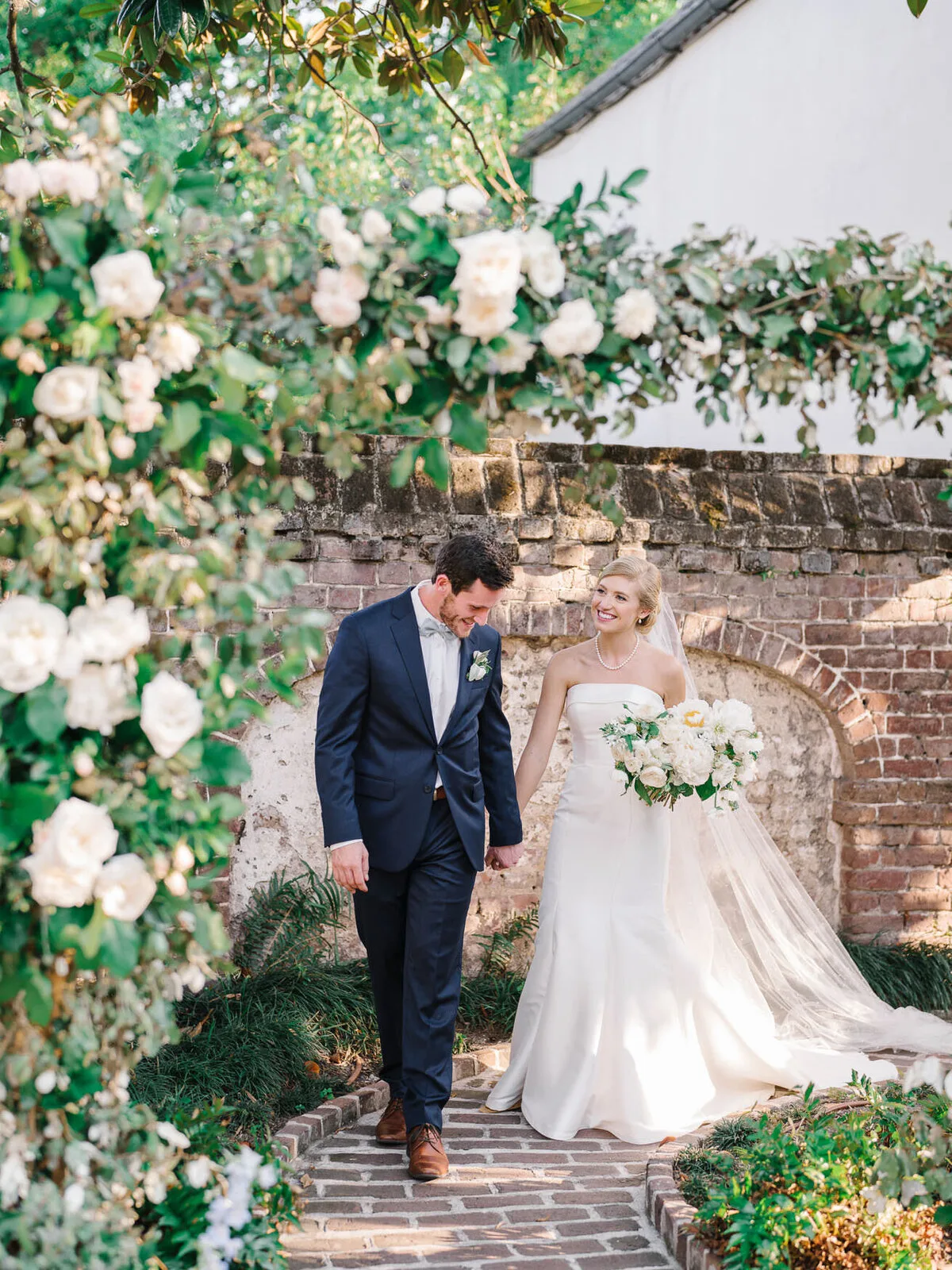 Couple walking past a brick wall covered in flowers at a Charleston SC wedding