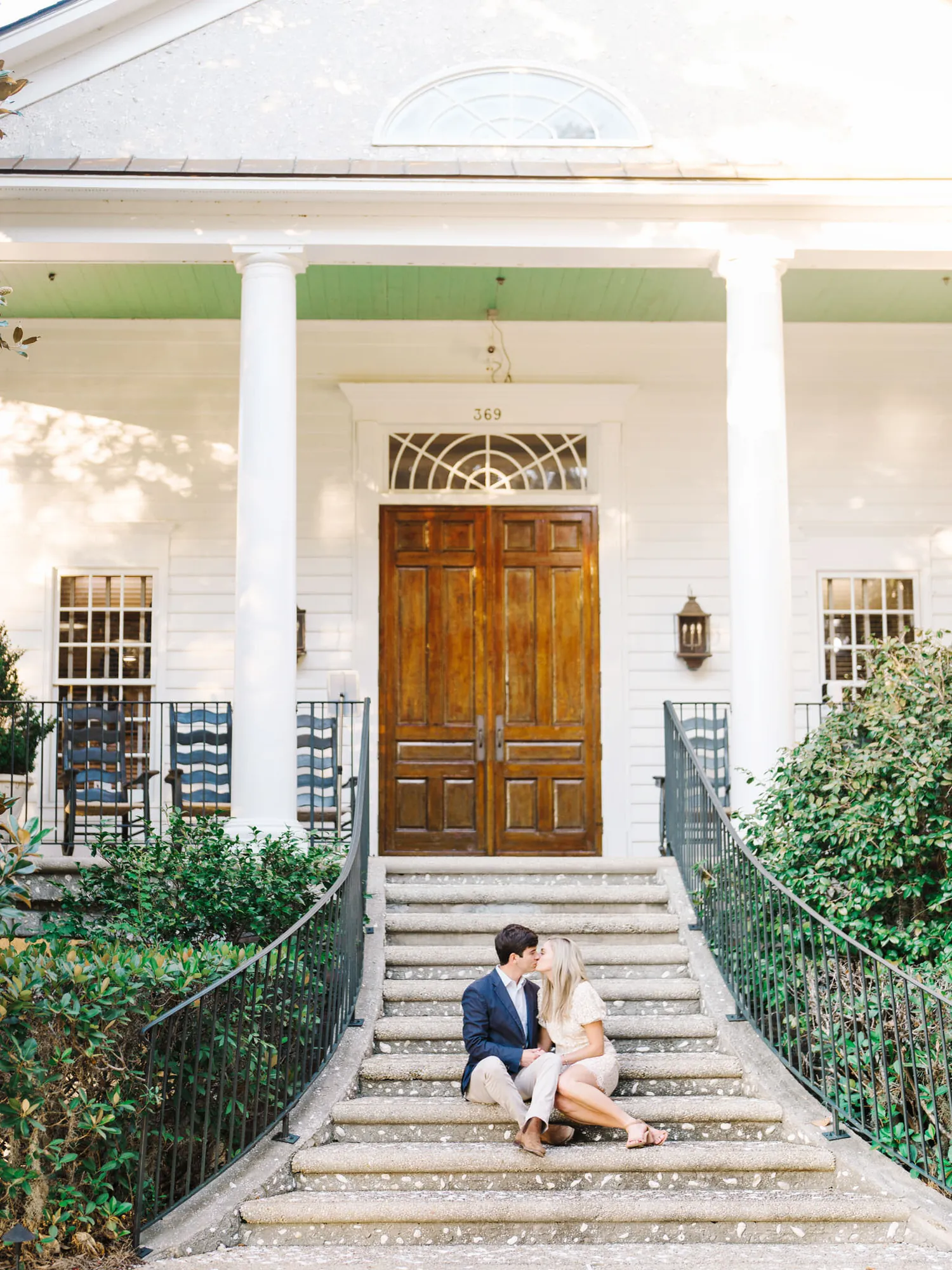Engaged couple sitting and kissing on the brick steps of a white columned plantation house with a wooden front door near Charleston