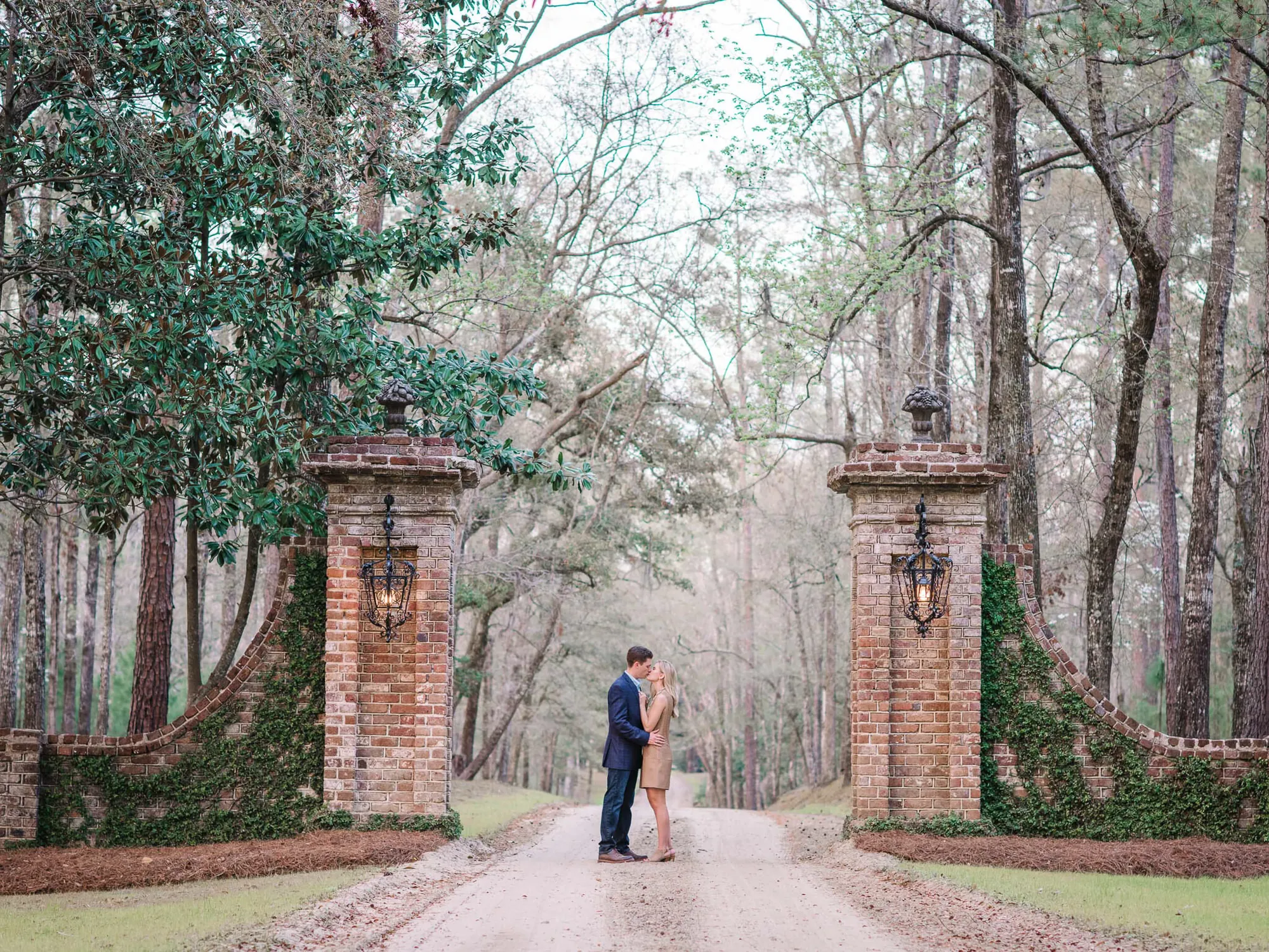 Engaged couple embracing on a long dirt drive between two brick gate posts at a South Carolina plantation