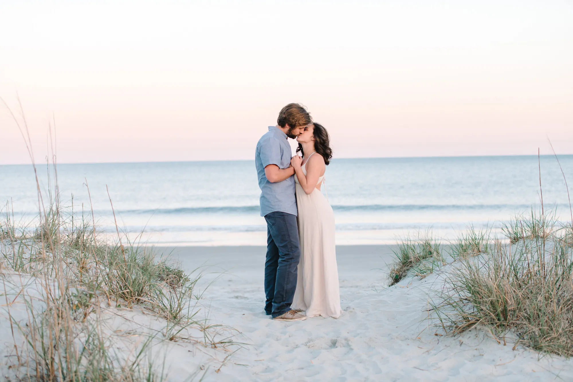 Engaged couple embracing in the sand dunes with sea oats and the ocean behind them at sunset in Myrtle Beach