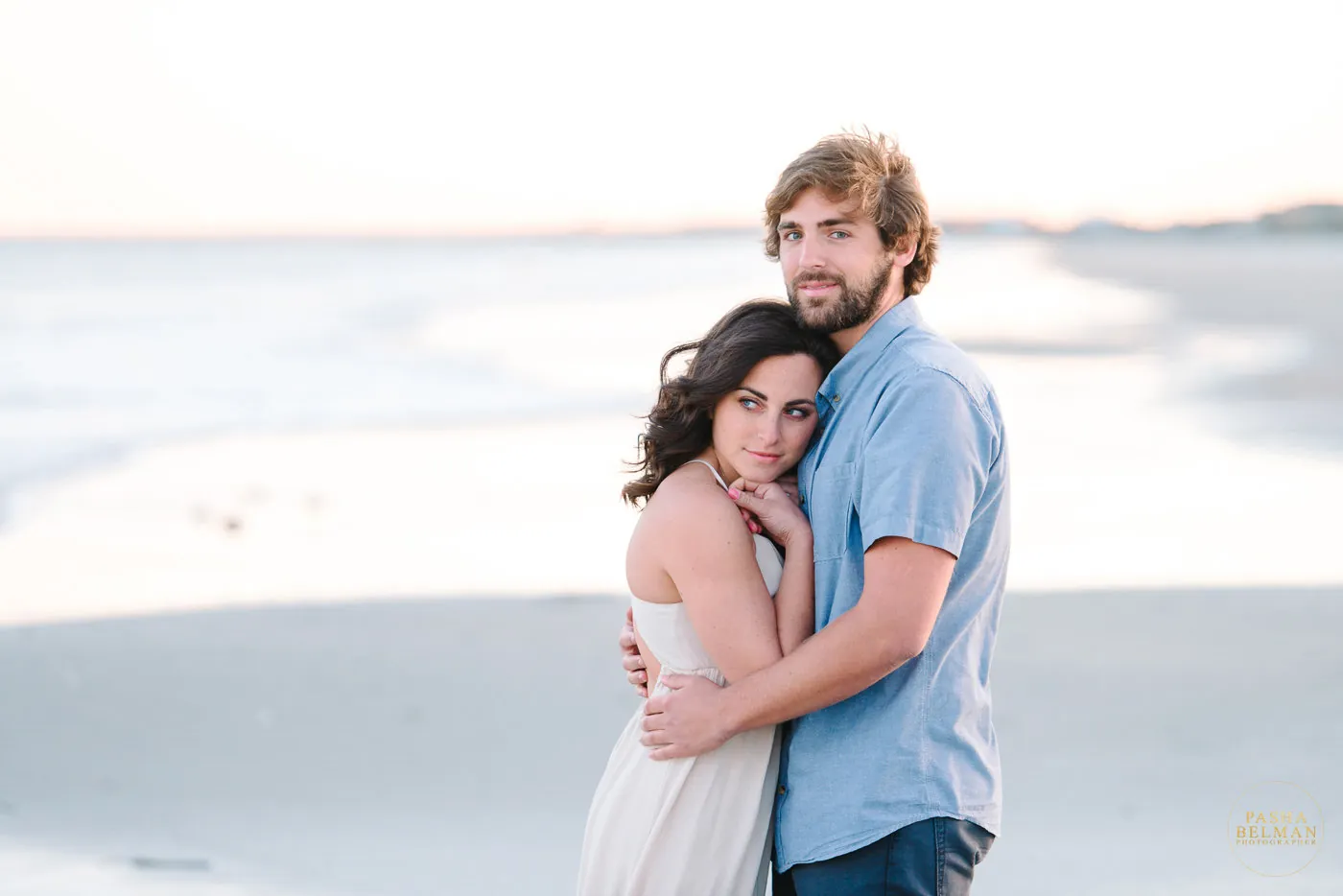 Engaged couple embracing on an empty beach at sunset, she in a cream dress and he in a blue button-up shirt, in Myrtle Beach