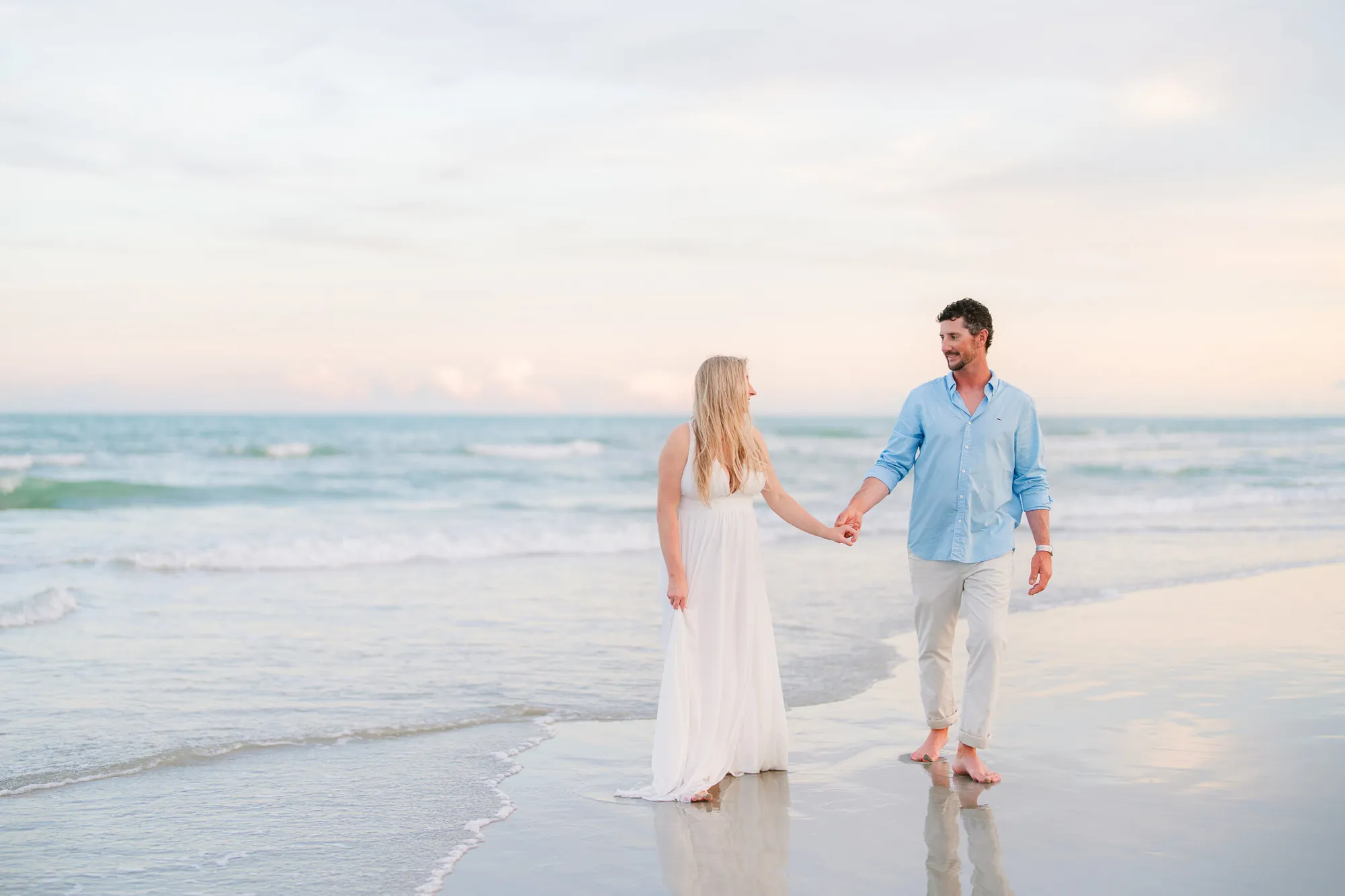 Engaged couple in a cream gown and blue shirt holding hands at the water edge under a pastel sunset in Myrtle Beach