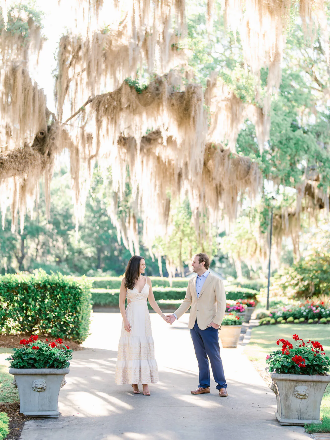 Engaged couple holding hands in a formal garden with red geranium planters and Spanish moss overhead in South Carolina