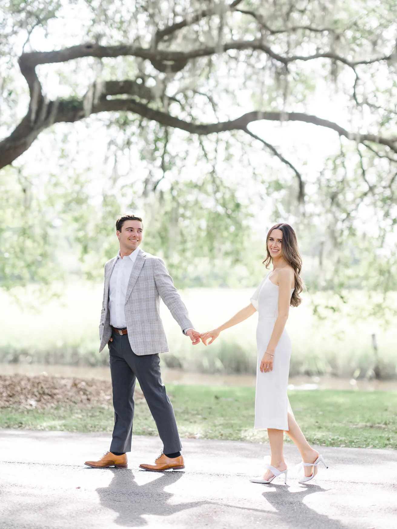 Engaged couple in a gray blazer and white dress holding hands on a quiet country road under a spreading live oak