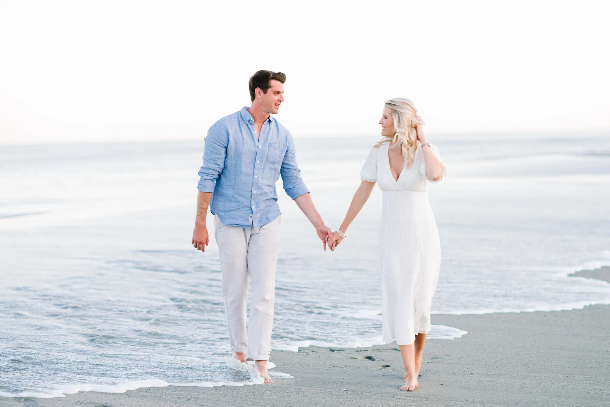 Engaged couple in blue linen and white holding hands while walking the shoreline at low tide in Myrtle Beach