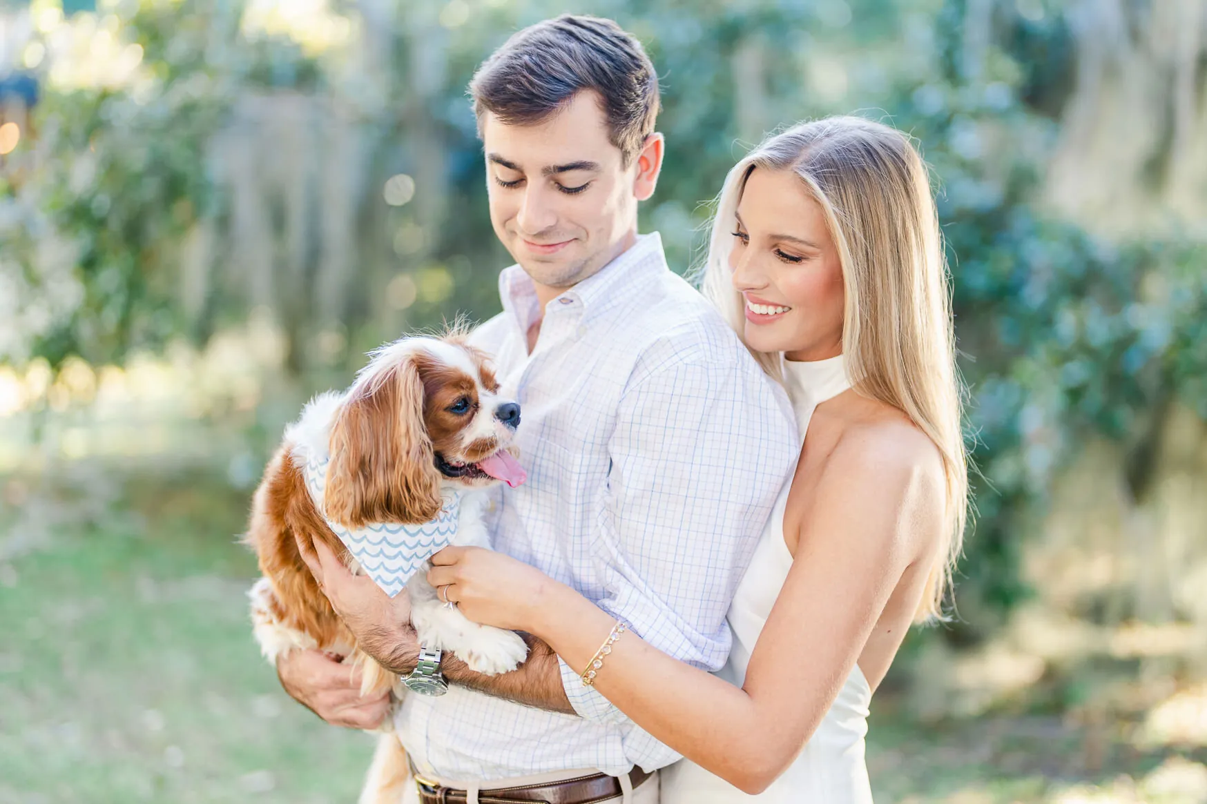 Engaged couple smiling while holding their Cavalier King Charles spaniel in a green garden setting on the Grand Strand