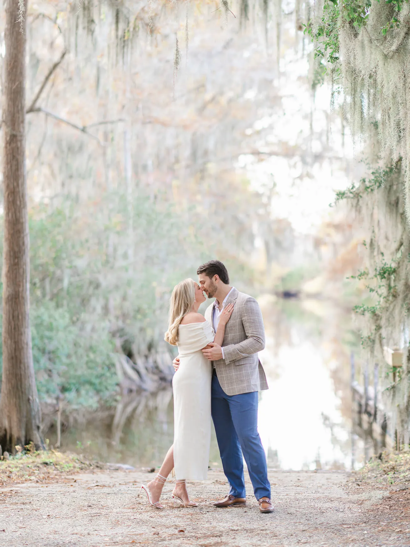 Engaged couple kissing on a sandy path beside a quiet creek framed by Spanish moss and cypress trees