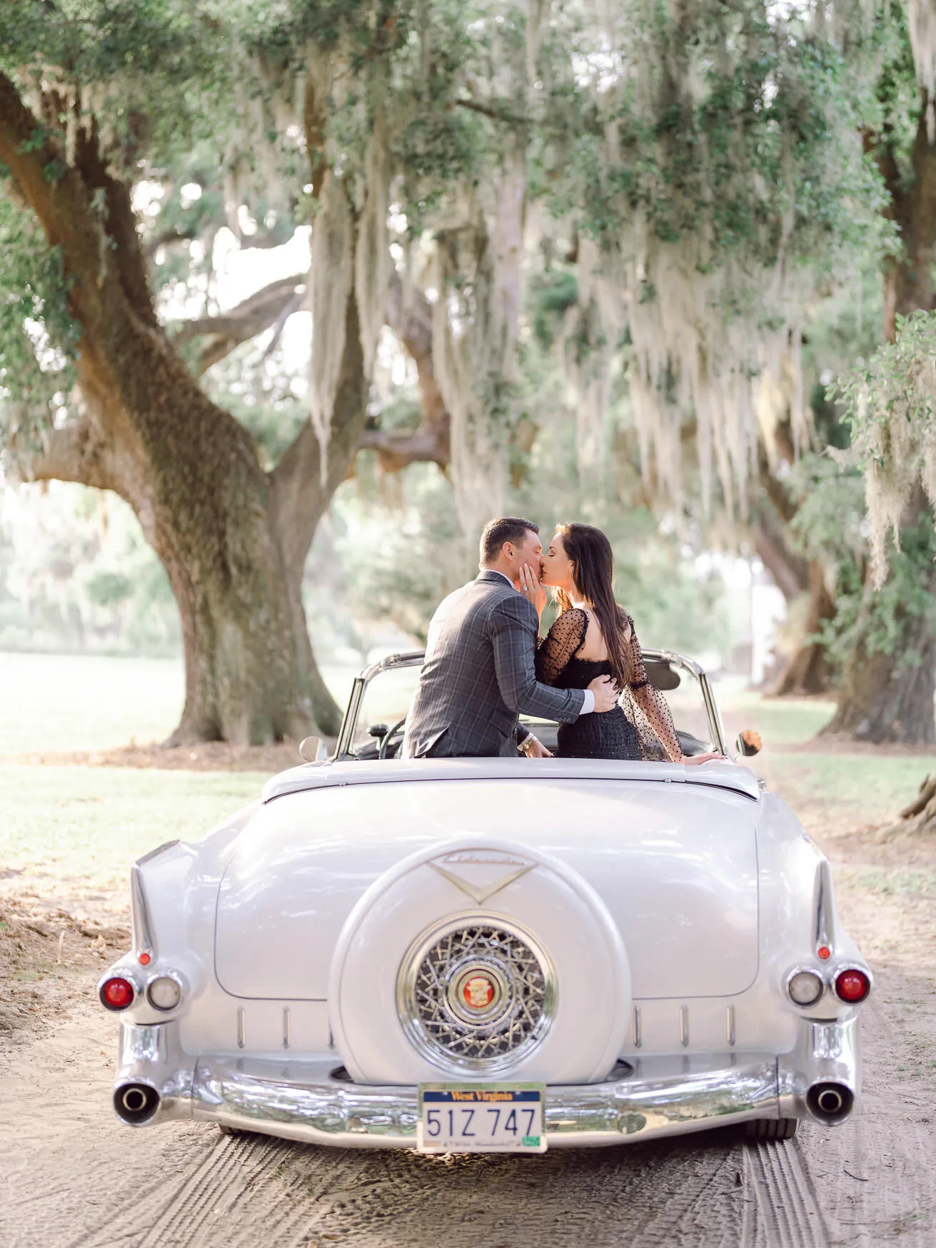 Engaged couple kissing in the back of a vintage white convertible parked under live oaks draped in Spanish moss