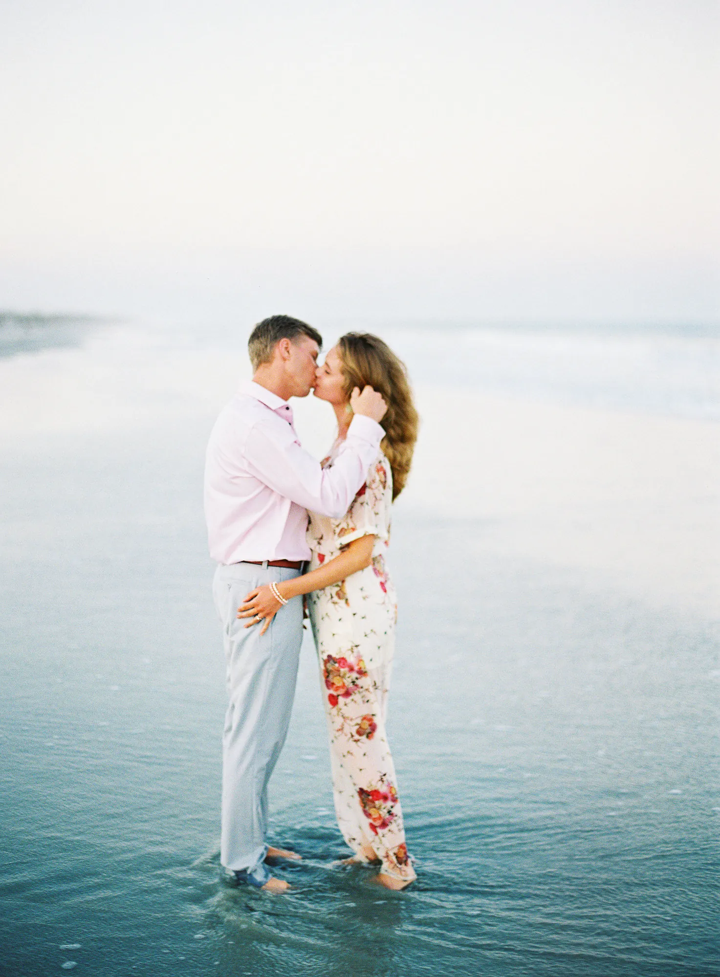 Engaged couple kissing in the shallow surf at dawn on a Myrtle Beach beach, she in a floral jumpsuit and he in a pink shirt