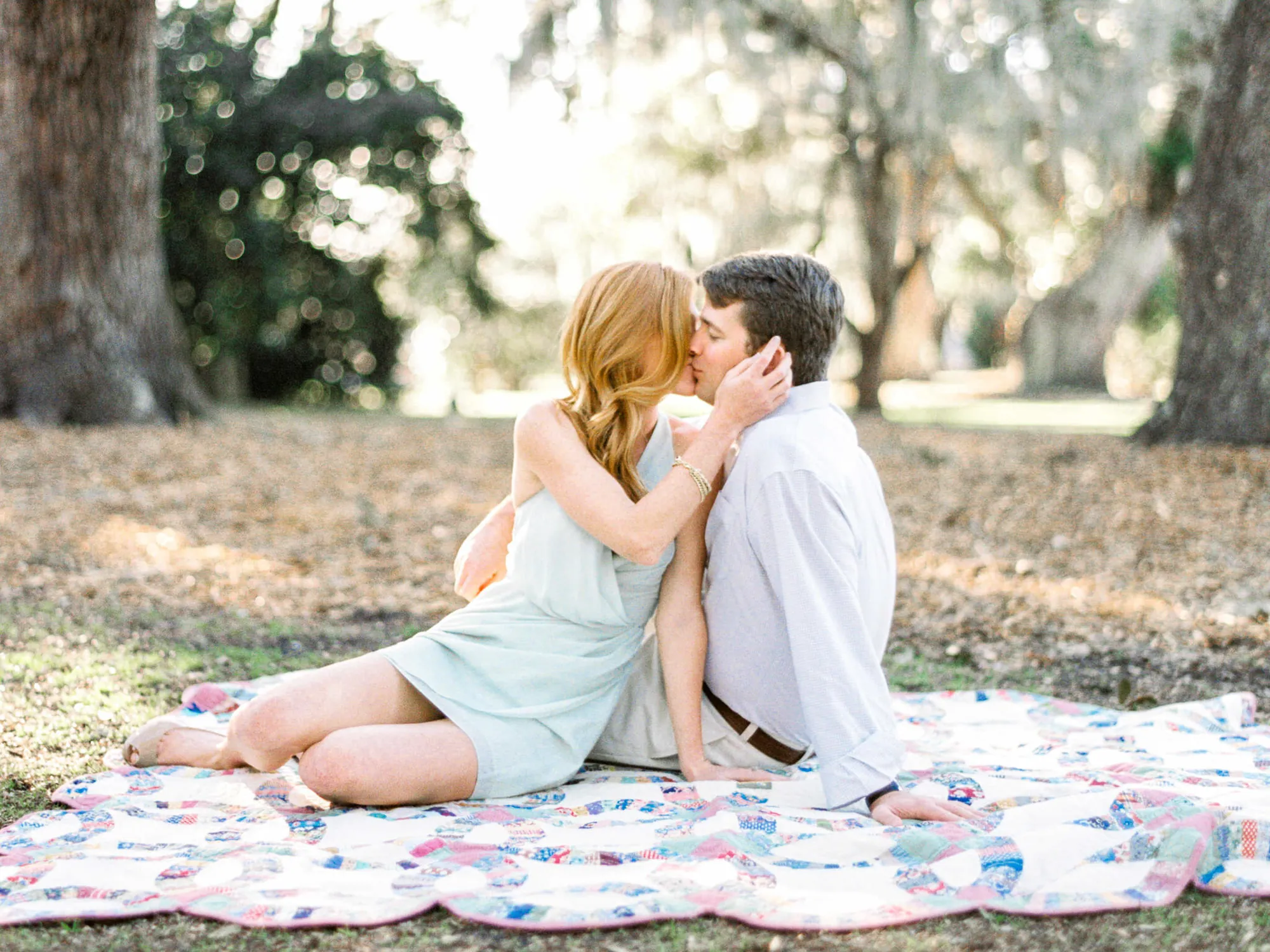 Engaged couple kissing while sitting on a patchwork quilt beneath live oak trees on the Grand Strand near Myrtle Beach