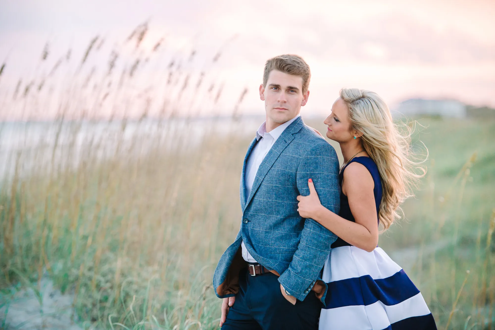 Engaged couple posing close together in tall golden sea oats at sunset, he in a blue blazer and she in a navy striped dress