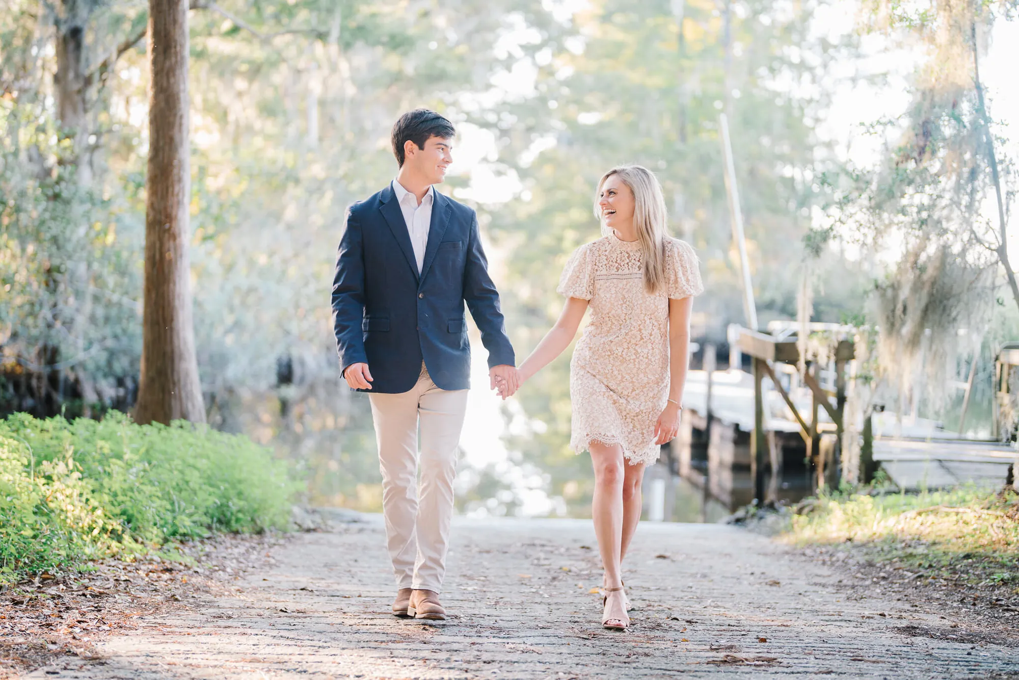 Engaged couple in a navy blazer and cream dress walking a brick path beside a boat dock and cypress trees on the Grand Strand