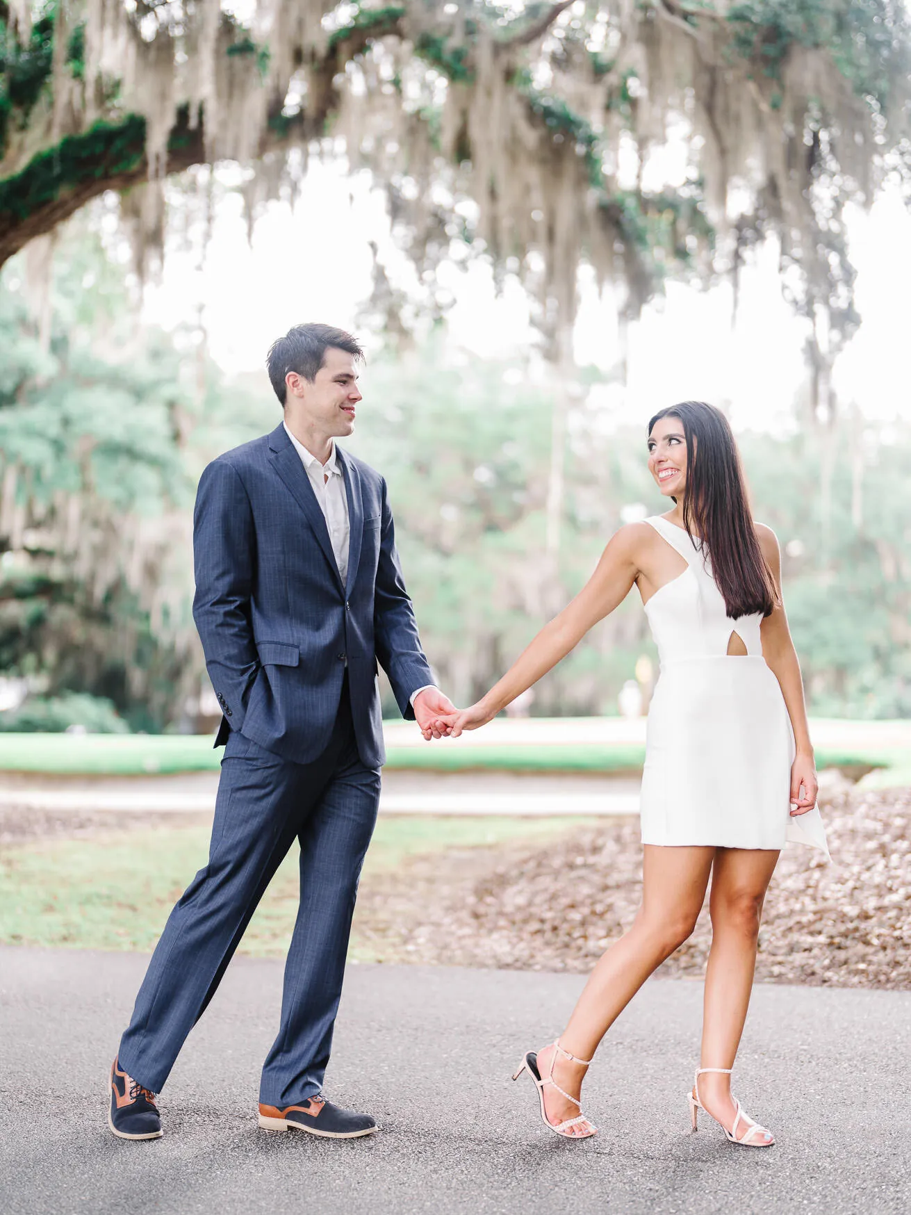 Engaged couple walking hand in hand under a live oak, he in a navy suit and she in a white cut-out dress, glancing back at each other