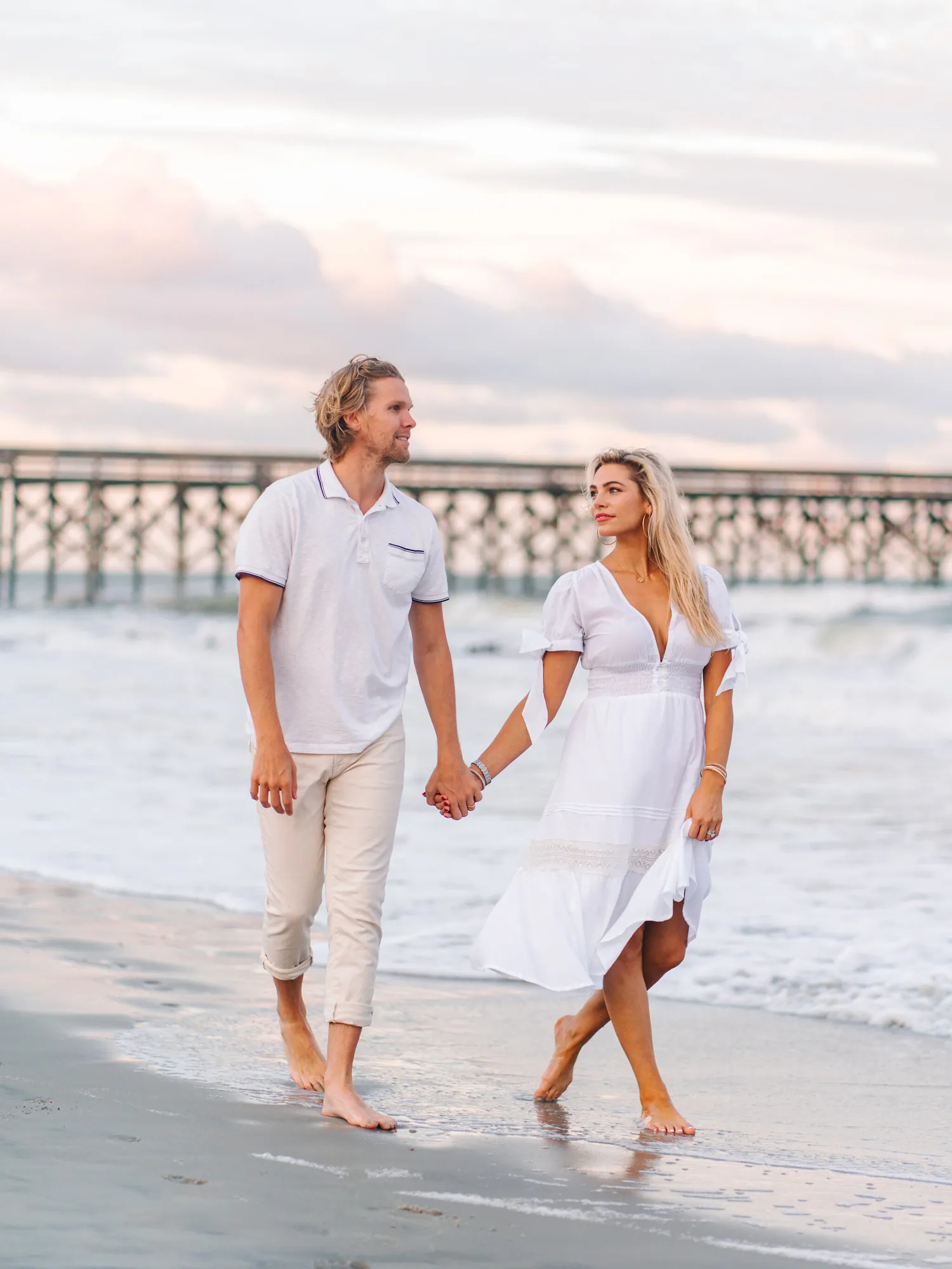 Engaged couple dressed in white holding hands and walking along the beach near a wooden pier at sunset in Myrtle Beach