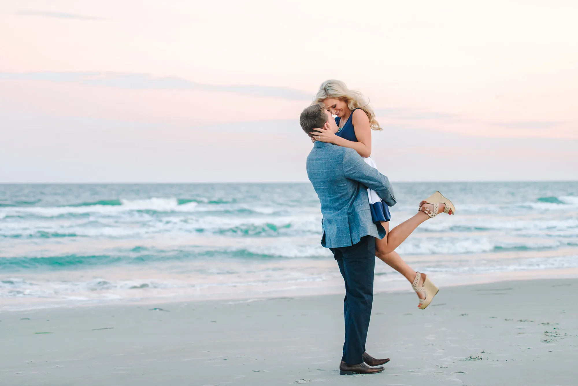 Man in a blue blazer lifting his fiancee off her feet and kissing her on a Myrtle Beach beach with turquoise waves behind them