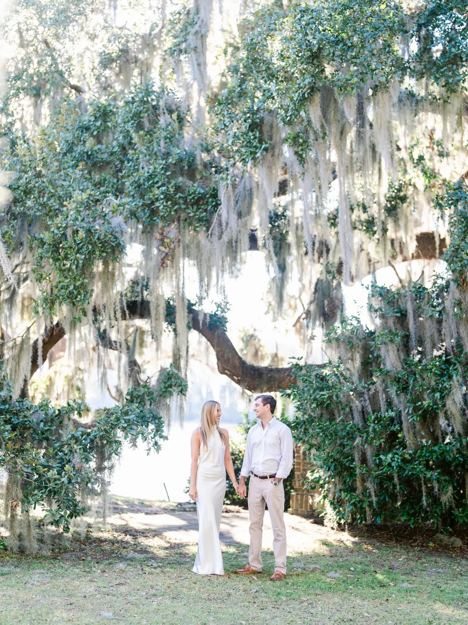 Engaged couple in white and khaki holding hands under a live oak dripping with Spanish moss at Wachesaw Plantation in Murrells Inlet