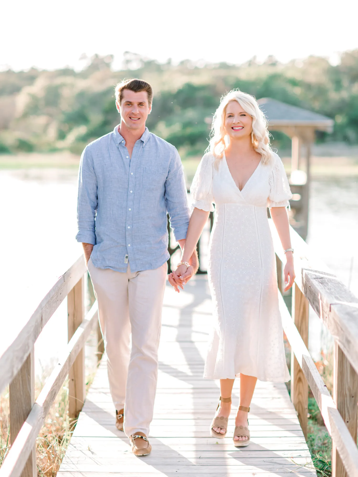 Engaged couple in blue linen and white walking hand in hand down a wooden boardwalk toward the marsh at Pawleys Island
