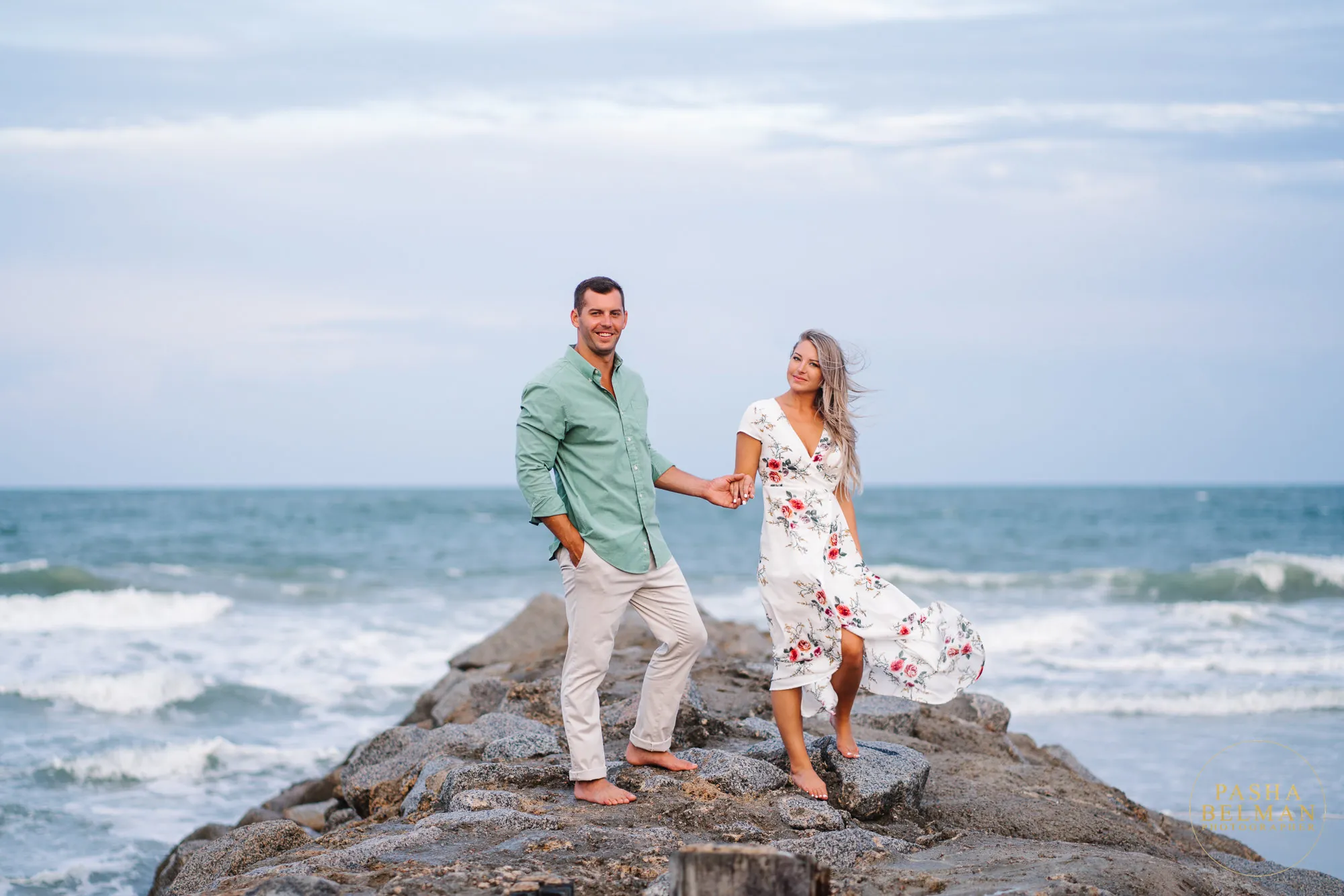 Engaged couple holding hands on a rocky jetty with ocean waves behind them at Pawleys Island, she in a floral dress