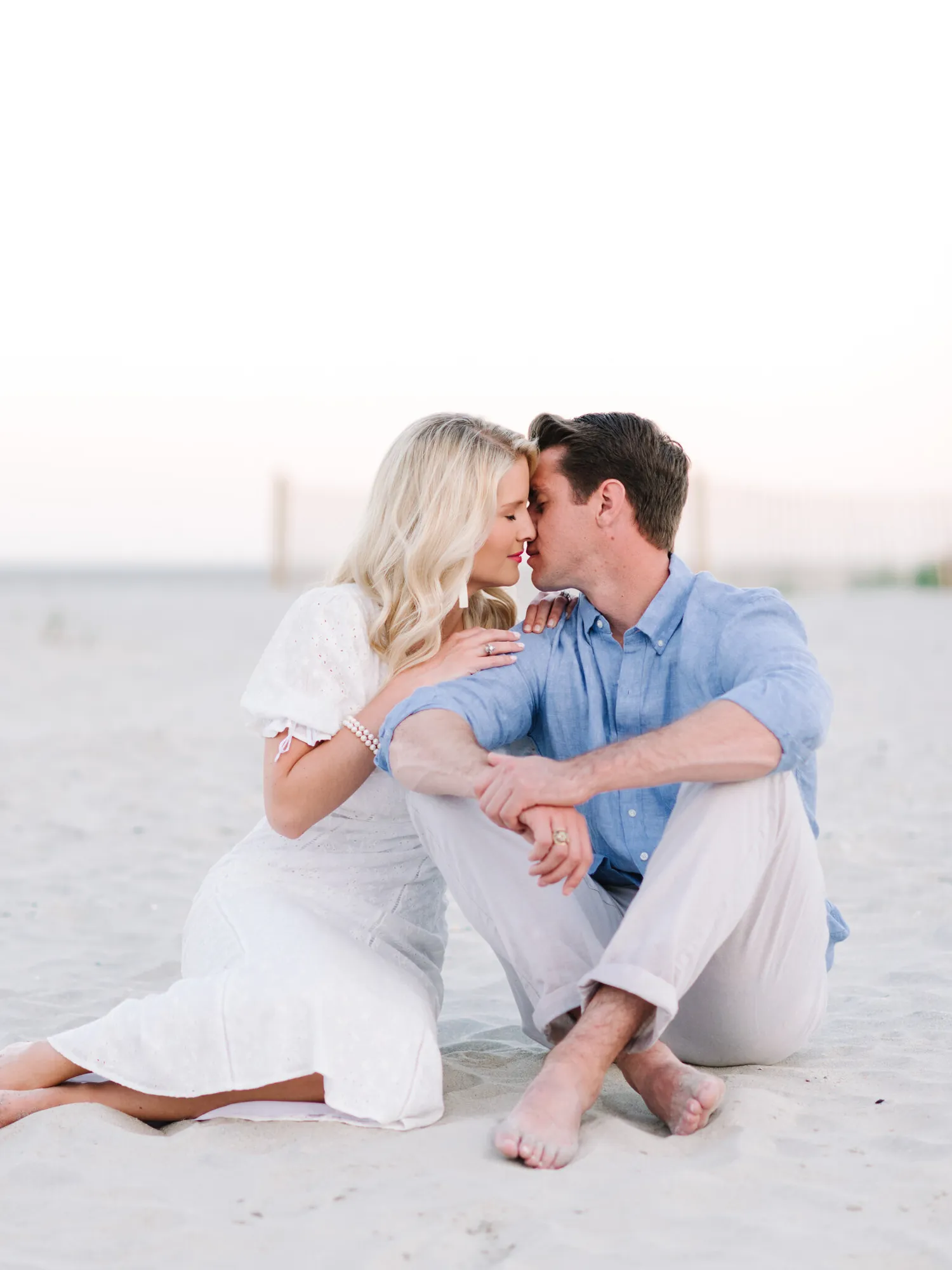 Engaged couple sitting close and nuzzling on the sand at Pawleys Island beach, she in a white puff sleeve dress and he in a blue shirt