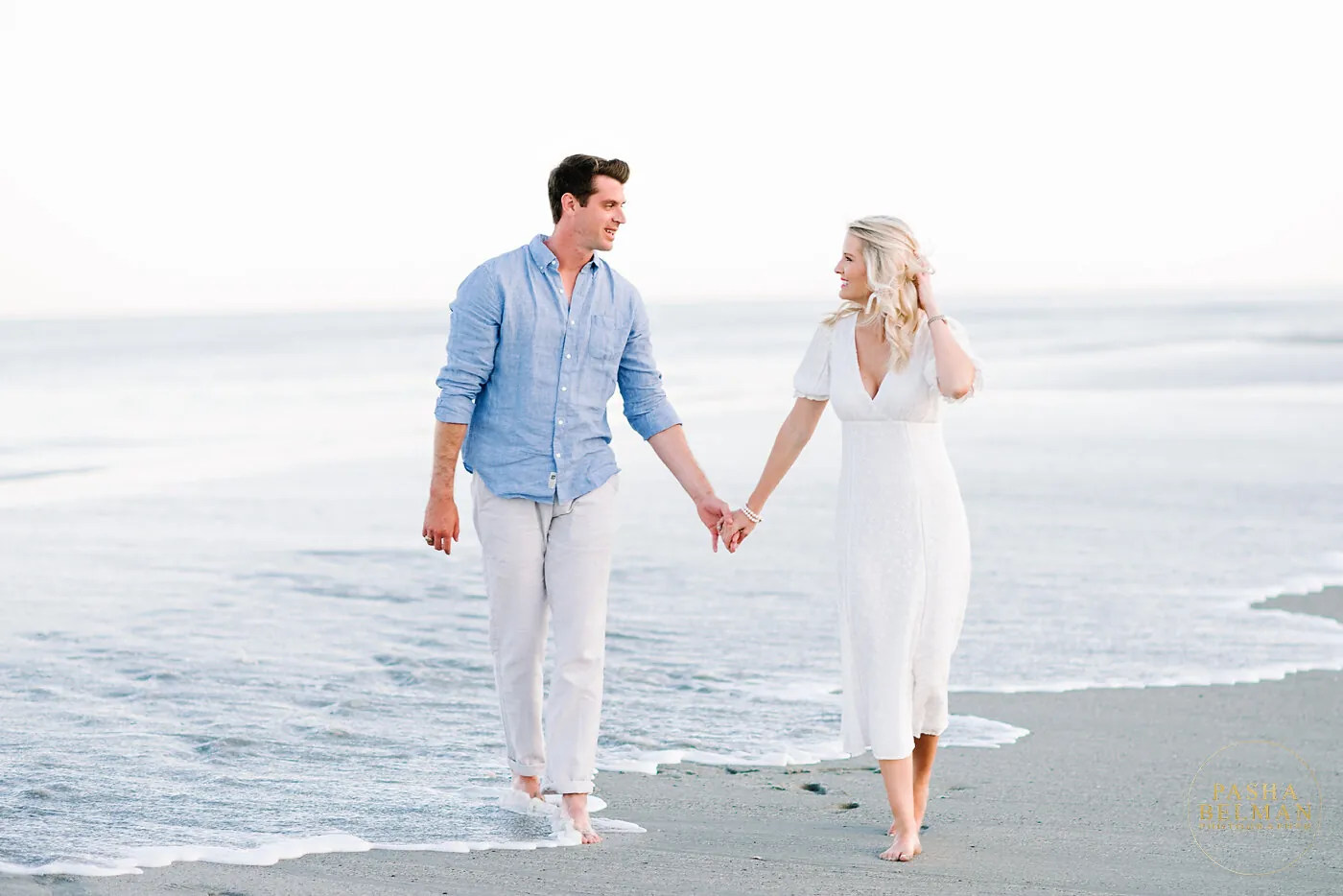 Engaged couple in light blue and white strolling the Pawleys Island shoreline holding hands at low tide
