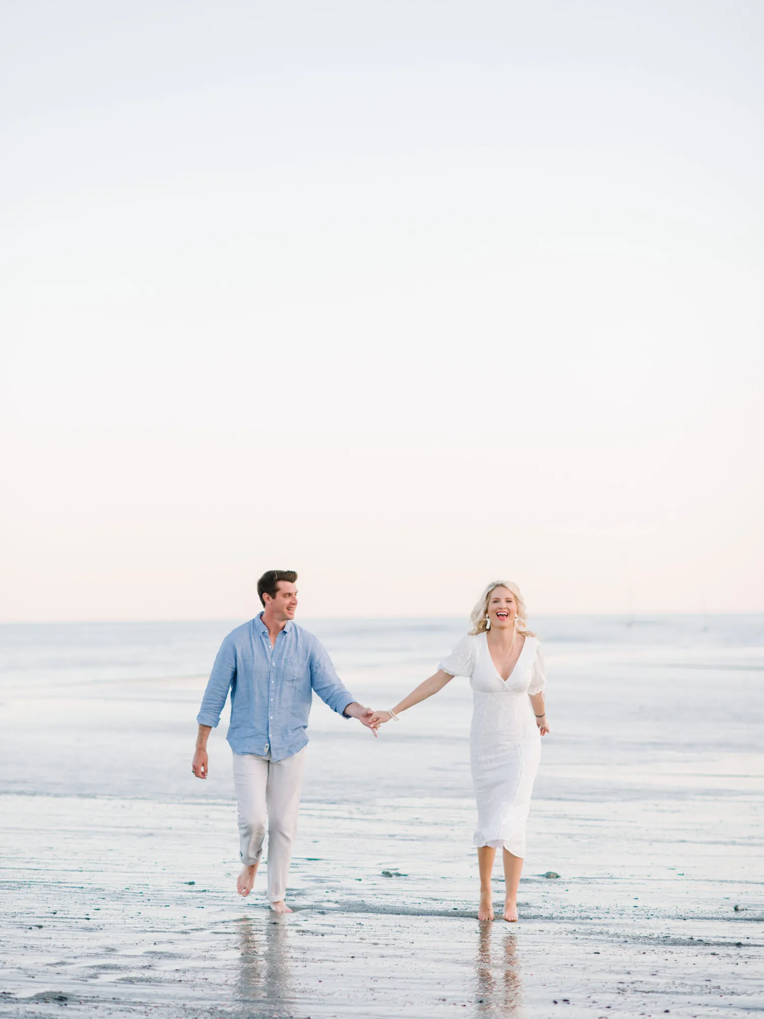 Engaged couple in light blue and white walking hand in hand through the shallow surf on Pawleys Island beach at sunrise