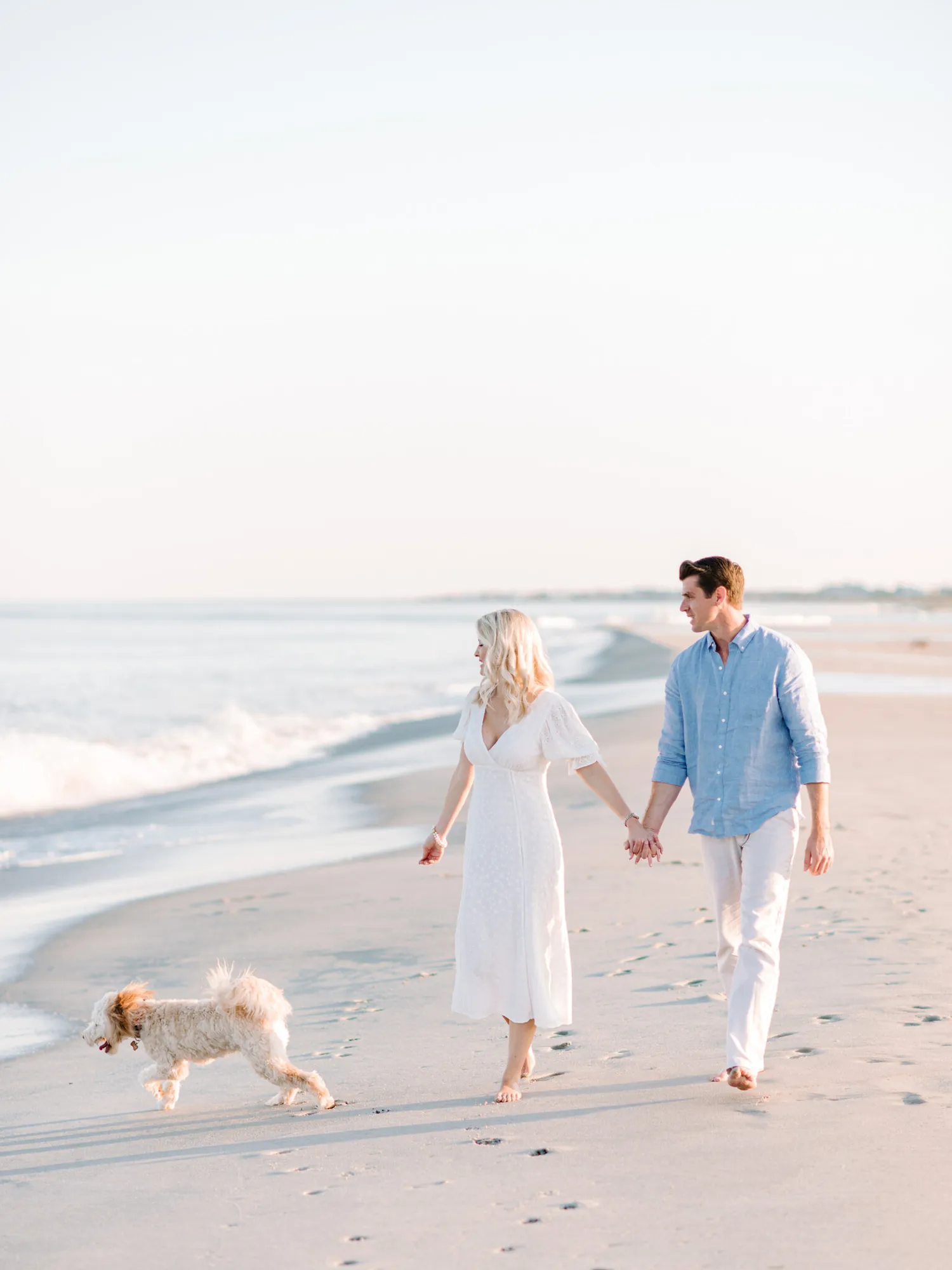 Engaged couple holding hands walking along the Pawleys Island shoreline with their small dog trotting beside them