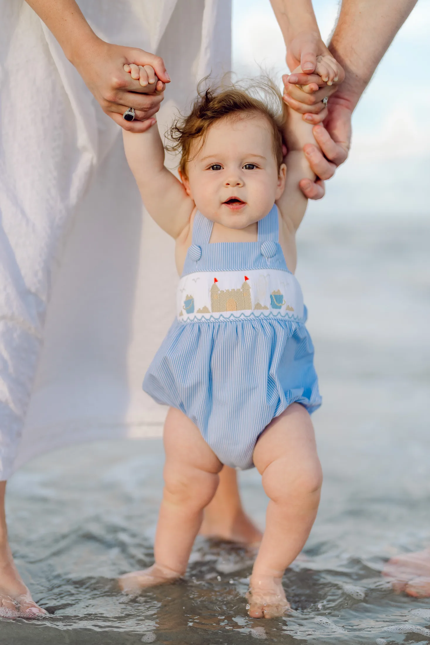 Baby in a blue sailboat romper learning to stand in the shoreline while parents hold his hands