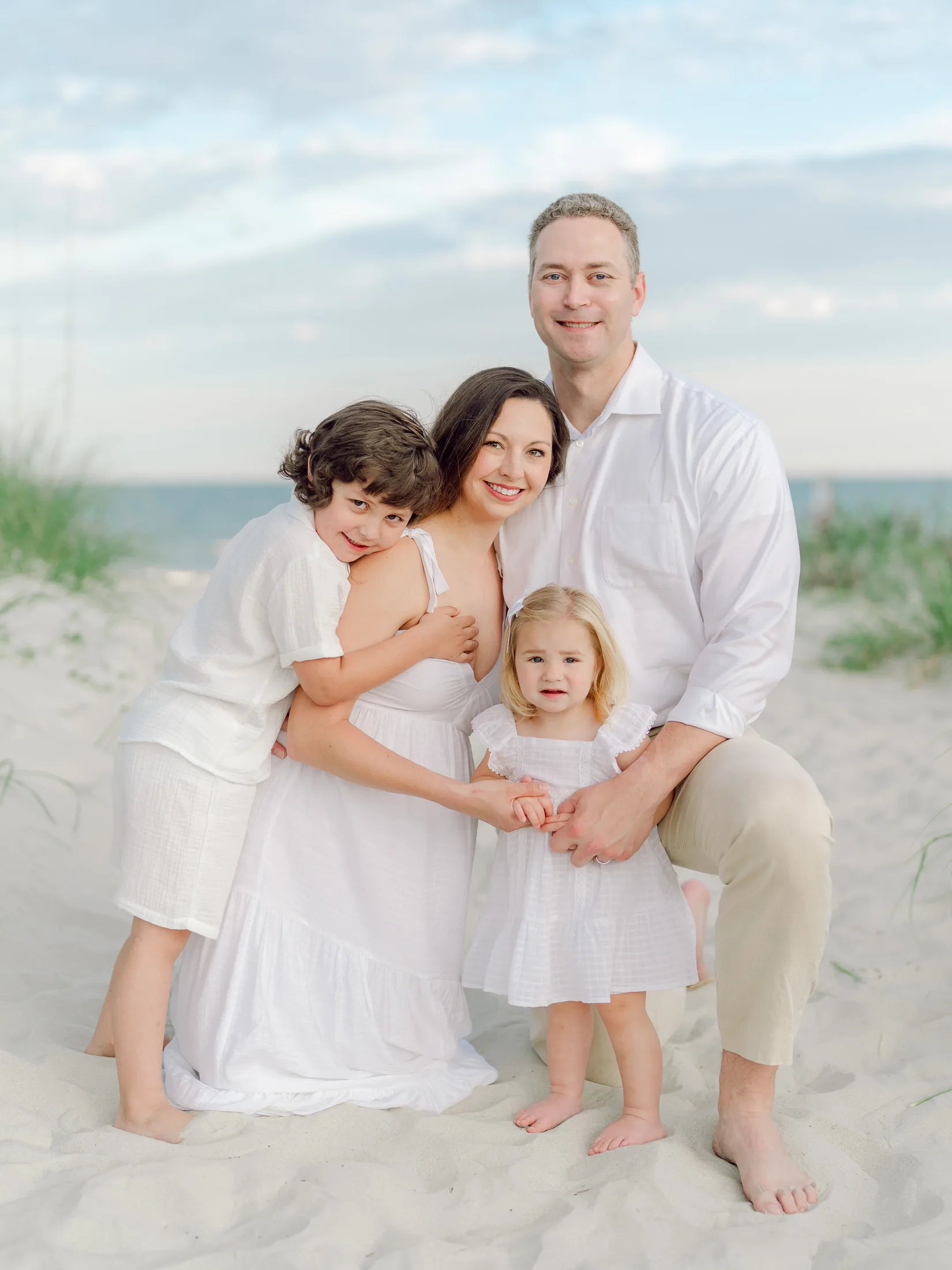 Family of four hugging on the sand dunes in white outfits at sunset