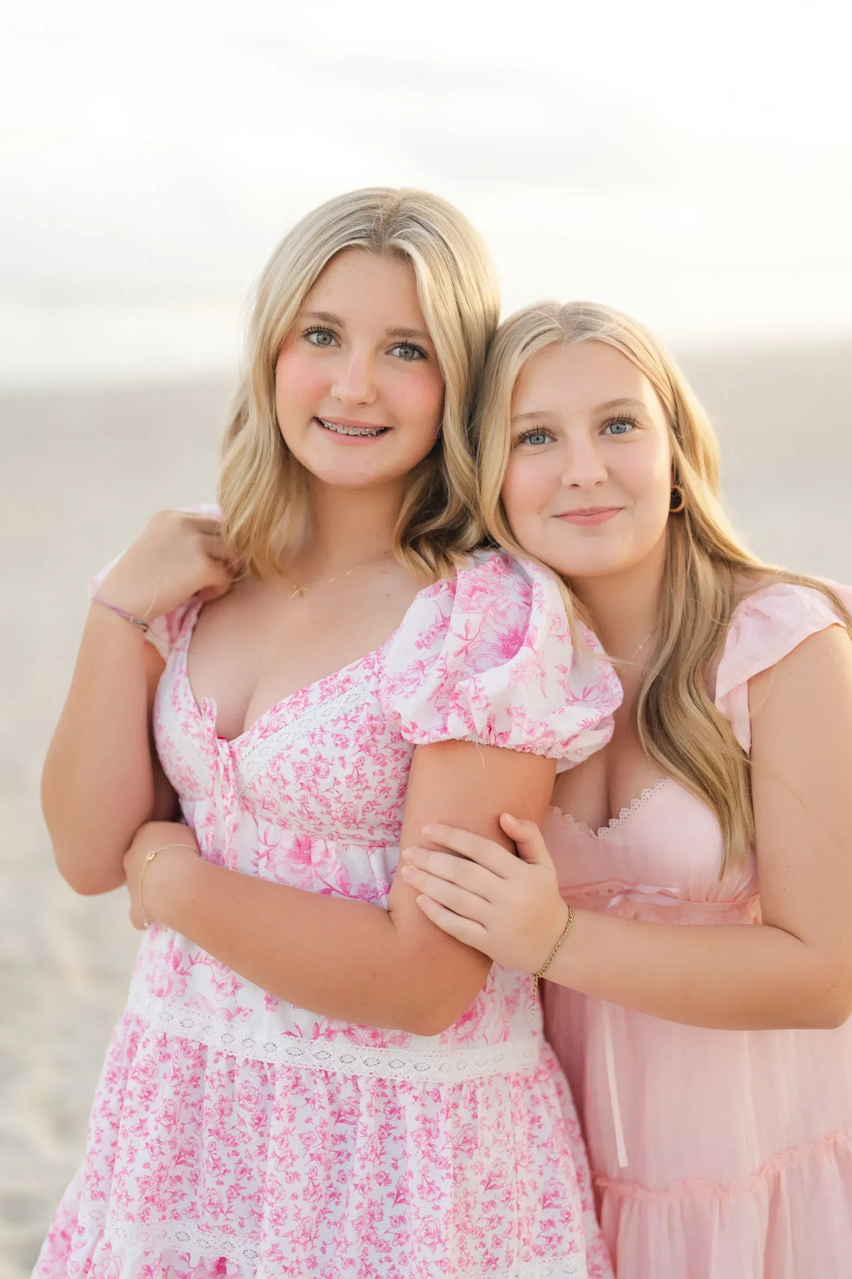 Two sisters in pink dresses hugging on the beach by Pasha Belman Photography