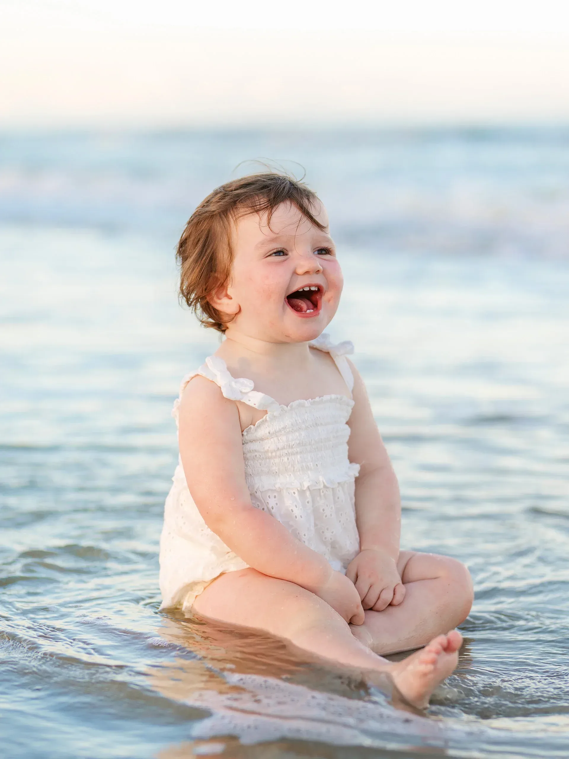 Baby girl laughing while sitting in the ocean shallows by Pasha Belman Photography