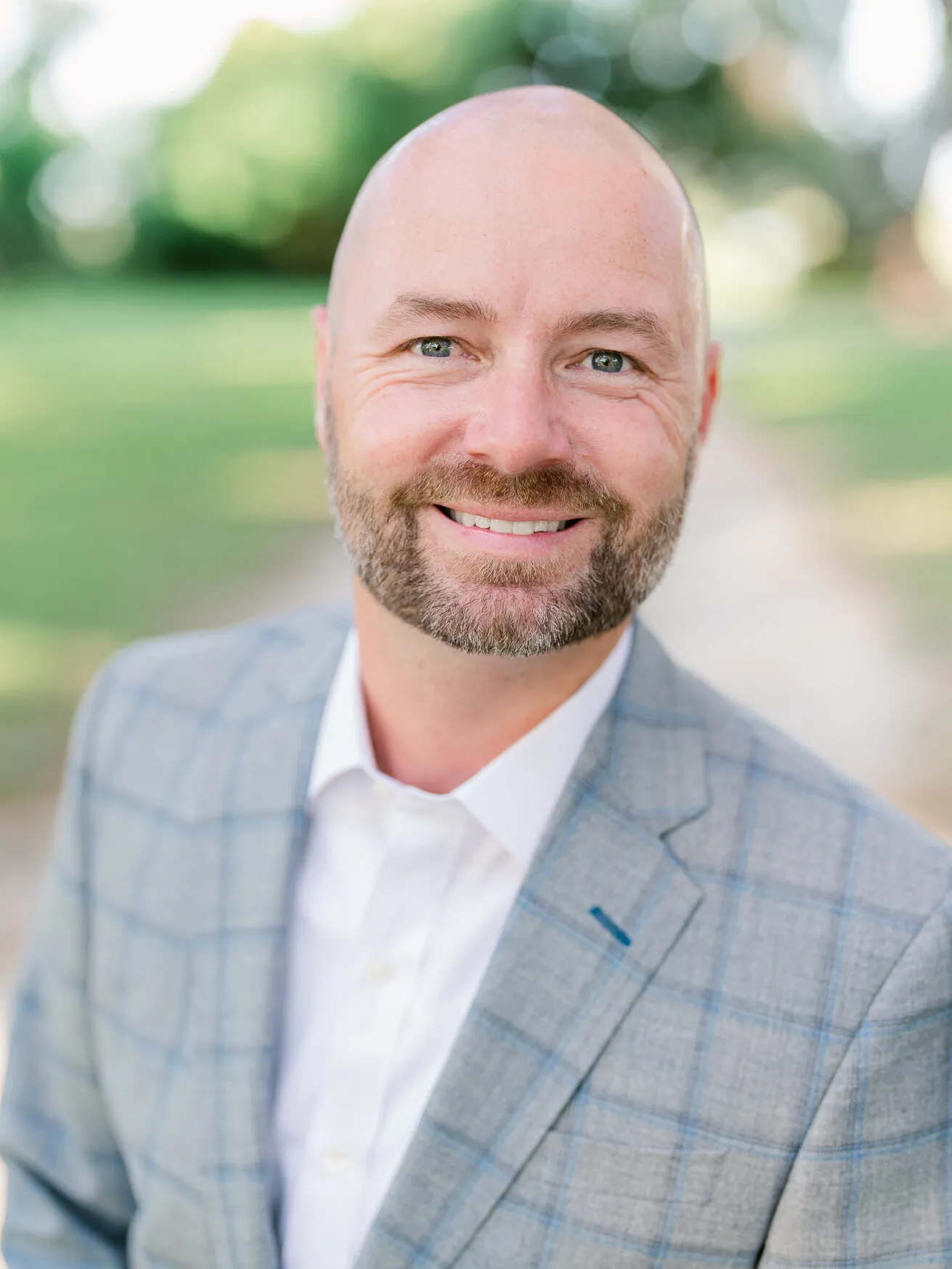 Professional headshot of a smiling bearded man in a light gray plaid blazer and white shirt, outdoors by Myrtle Beach headshot photographer Pasha Belman