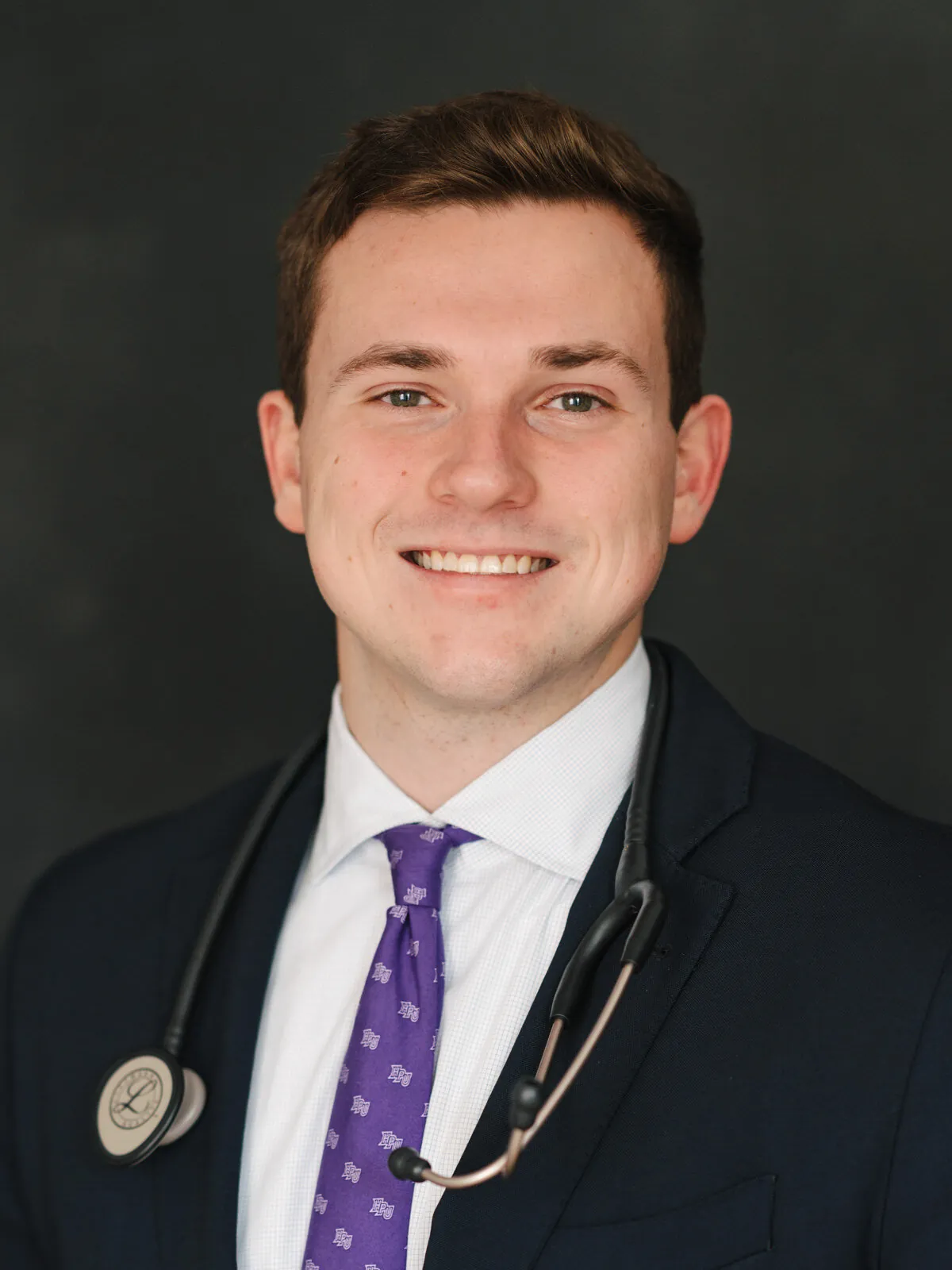 Professional headshot of a young doctor in a navy suit, white shirt and purple tie with a stethoscope around his neck, on a dark background by Myrtle Beach headshot photographer Pasha Belman
