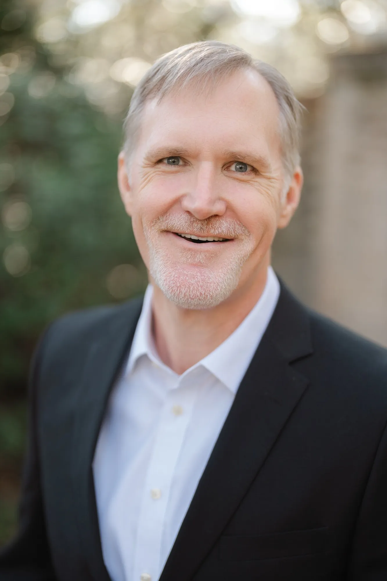 Professional headshot of a smiling man with graying hair in a black blazer and white open-collar shirt beside a stone wall by Myrtle Beach headshot photographer Pasha Belman