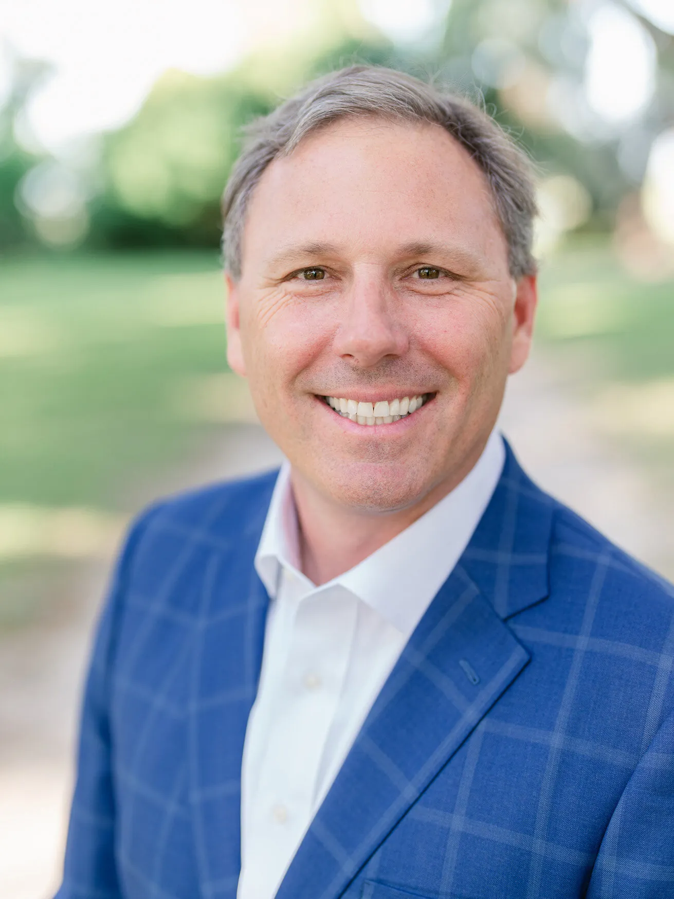 Professional headshot of a smiling man with gray-brown hair in a bright blue plaid blazer and white shirt, outdoors by Myrtle Beach headshot photographer Pasha Belman