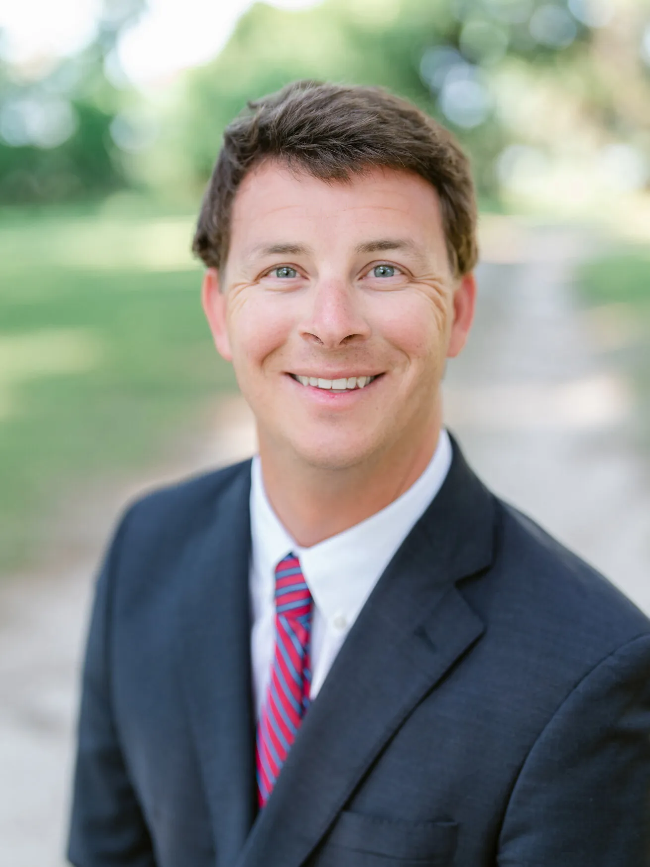 Professional headshot of a smiling man in a dark suit, white shirt and pink striped tie, outdoors by Myrtle Beach headshot photographer Pasha Belman
