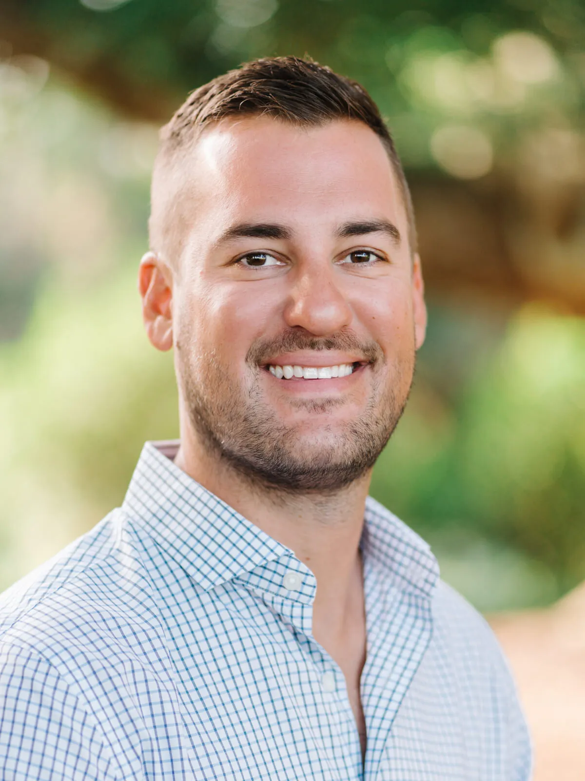 Professional headshot of a smiling man with short dark hair in a light blue checked shirt, outdoors by Myrtle Beach headshot photographer Pasha Belman