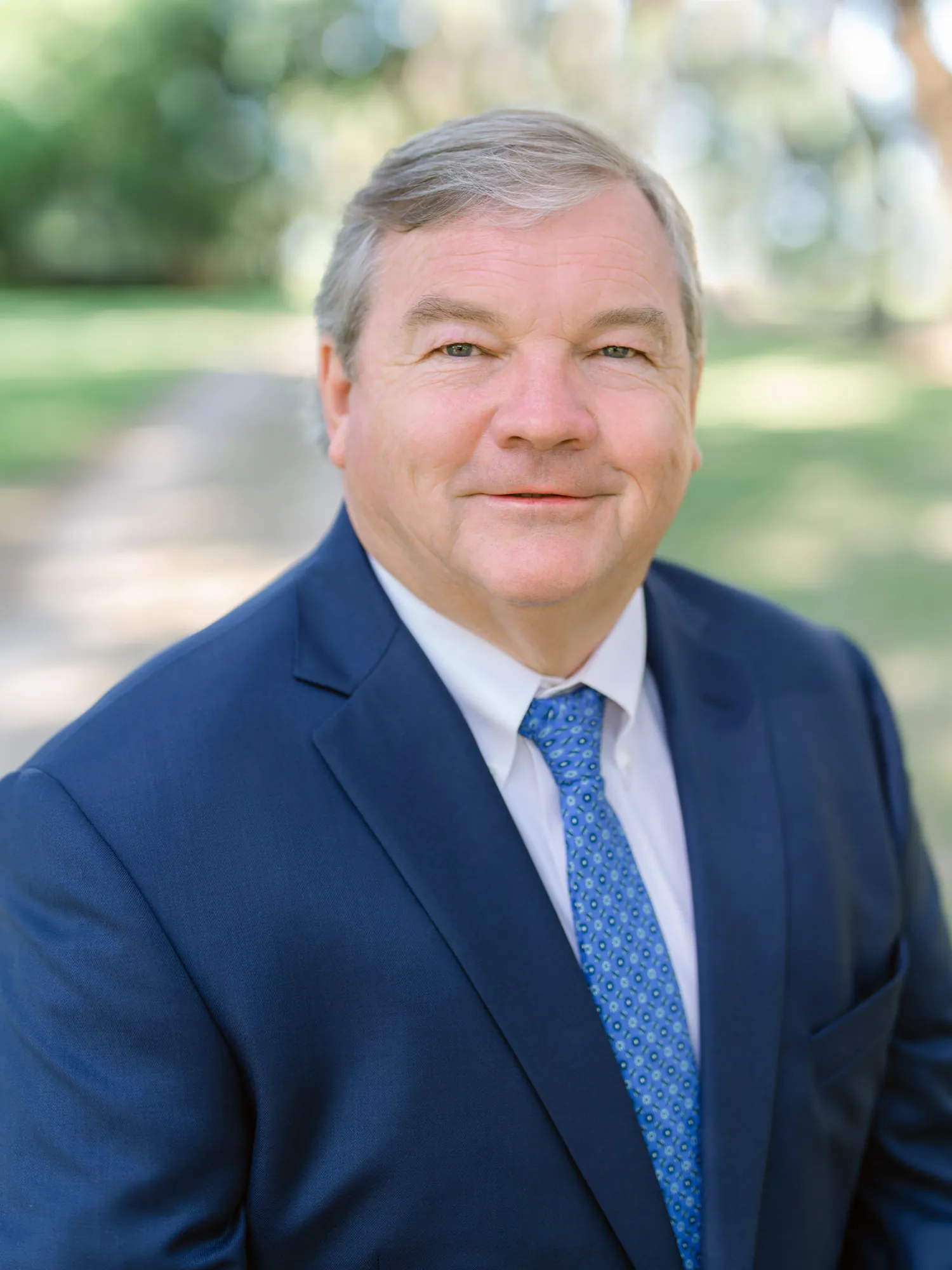 Professional headshot of a man in a navy suit with a blue patterned tie, outdoors in a park by Myrtle Beach headshot photographer Pasha Belman