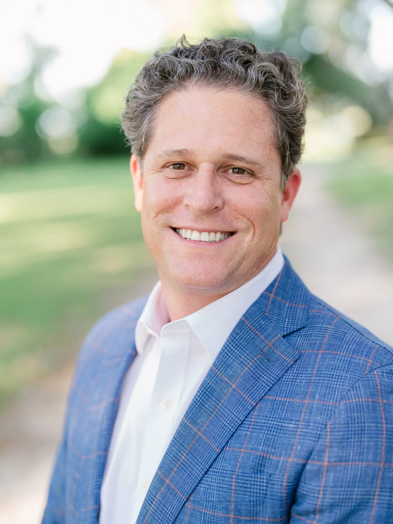 Professional headshot of a smiling man with curly gray-brown hair in a blue plaid blazer and white shirt, outdoors by Myrtle Beach headshot photographer Pasha Belman